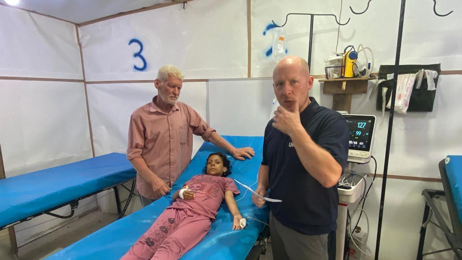 Mr Sears treating a girl called in Gaza. The young girl is lying on a blue bed and is hooked up to a machine. Another man is standing next to her with his hand just above her head. She looks unwell and both Mr Sears and the other man appear worried. 