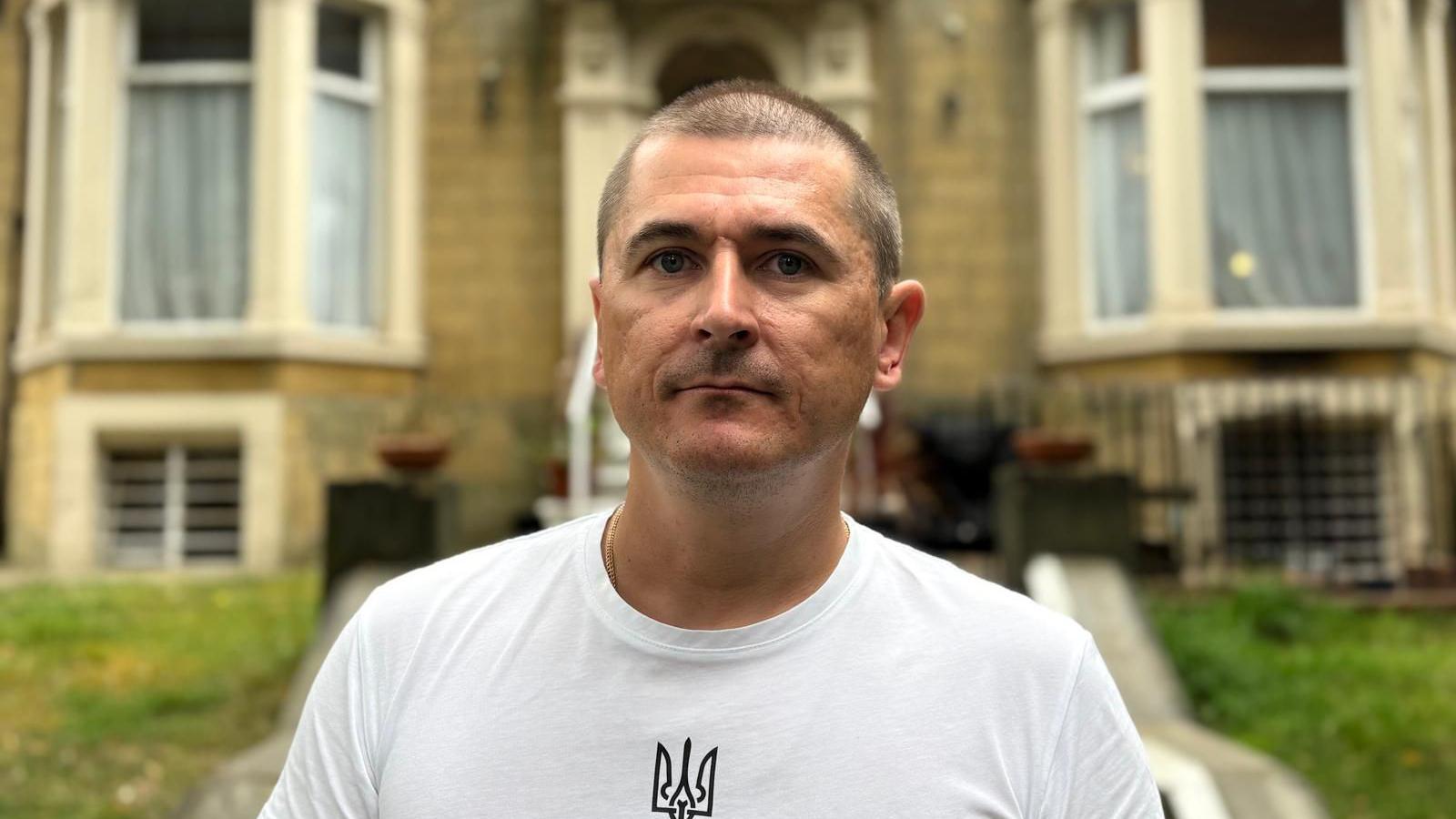 A man in a white t-shirt and short hair looks at the camera. Behind him is the Leeds Ukrainian Community Association building.