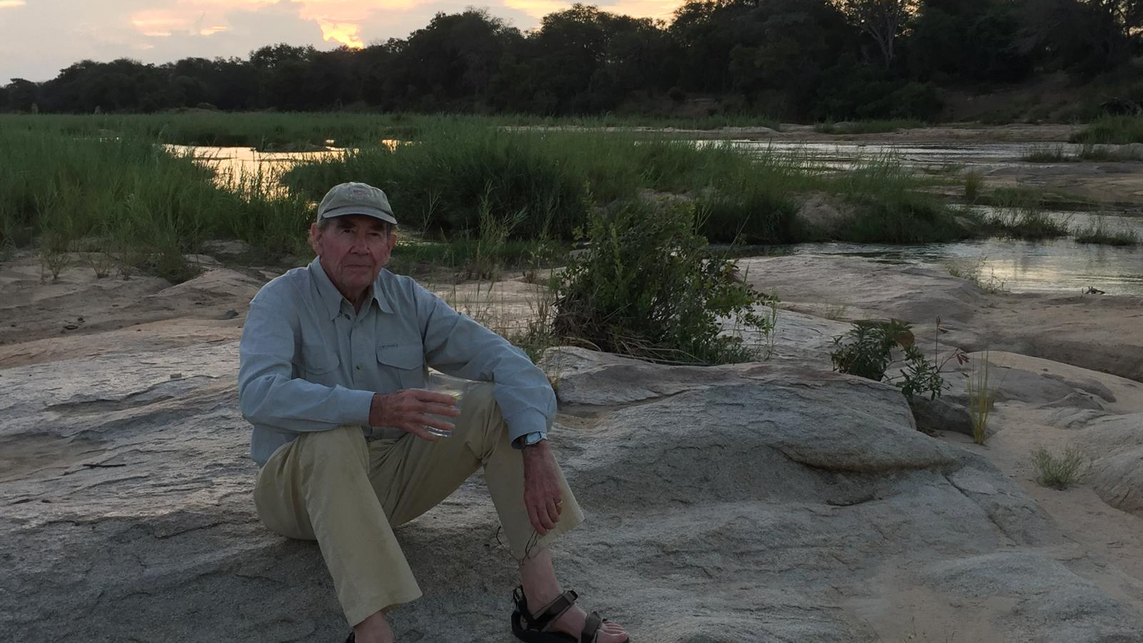 Sir Terence sits on a rocky area in light beige trousers and a blue shirt. Behind him is an orange and purple sunset.