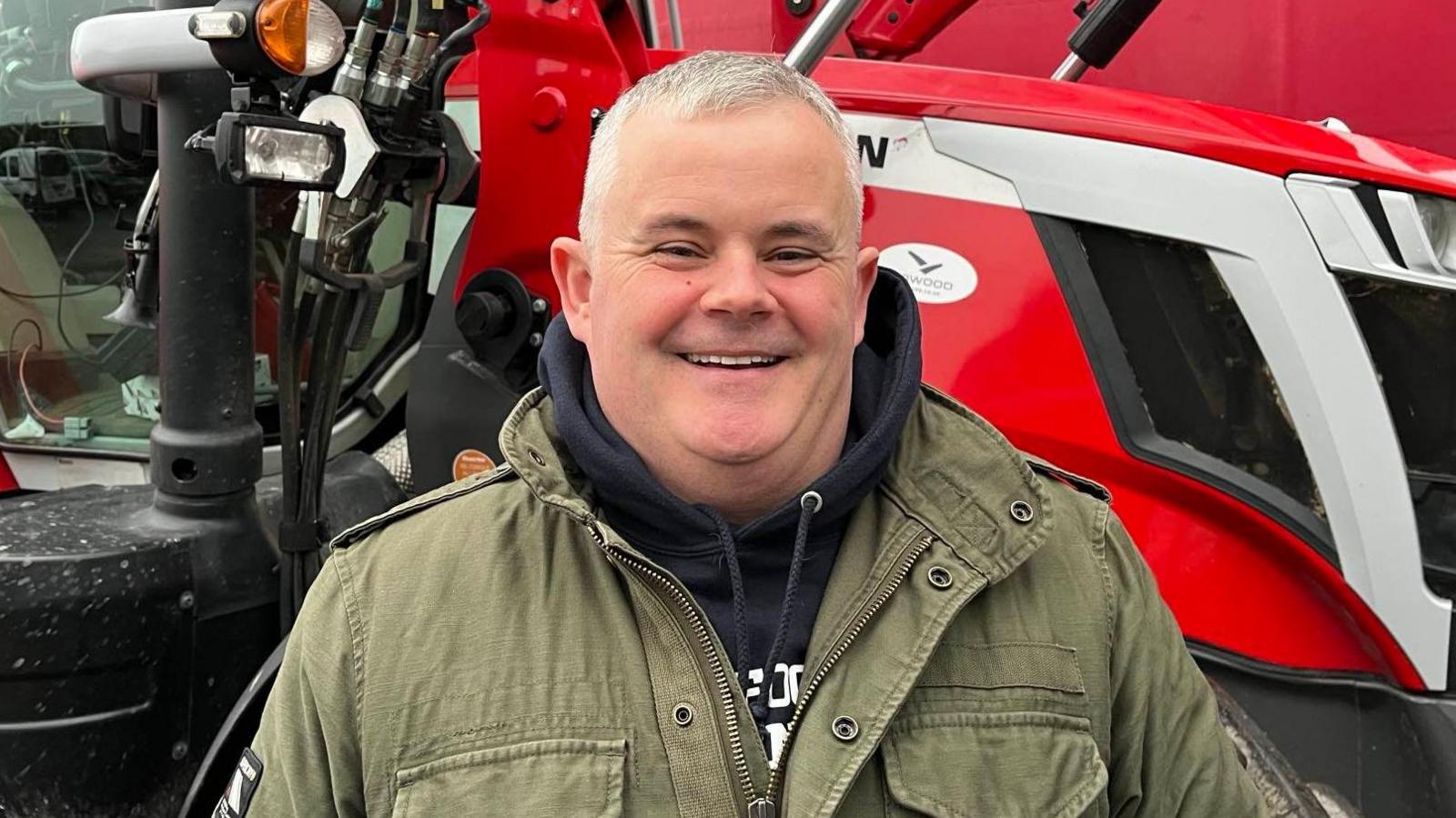 A man with short white hair. He is wearing a dark hoodie under a green coat and is standing in front of a red tractor.