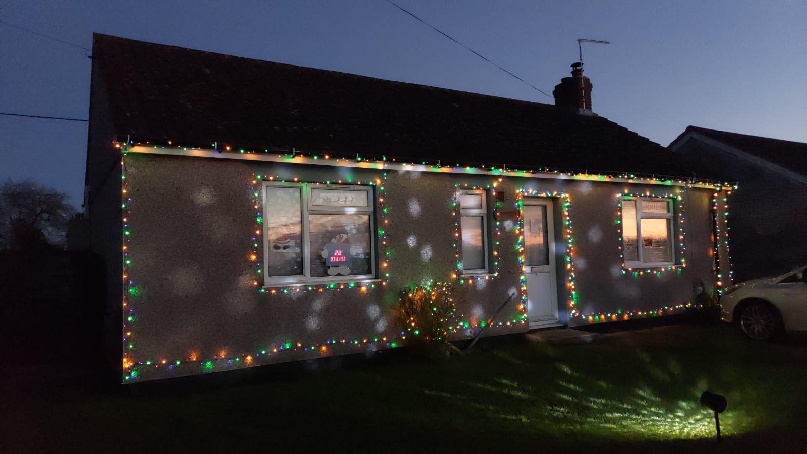 A bungalow is pictured at night with multi-coloured Christmas lights around it. Other lights are projected onto the garden grass in front of the home. 