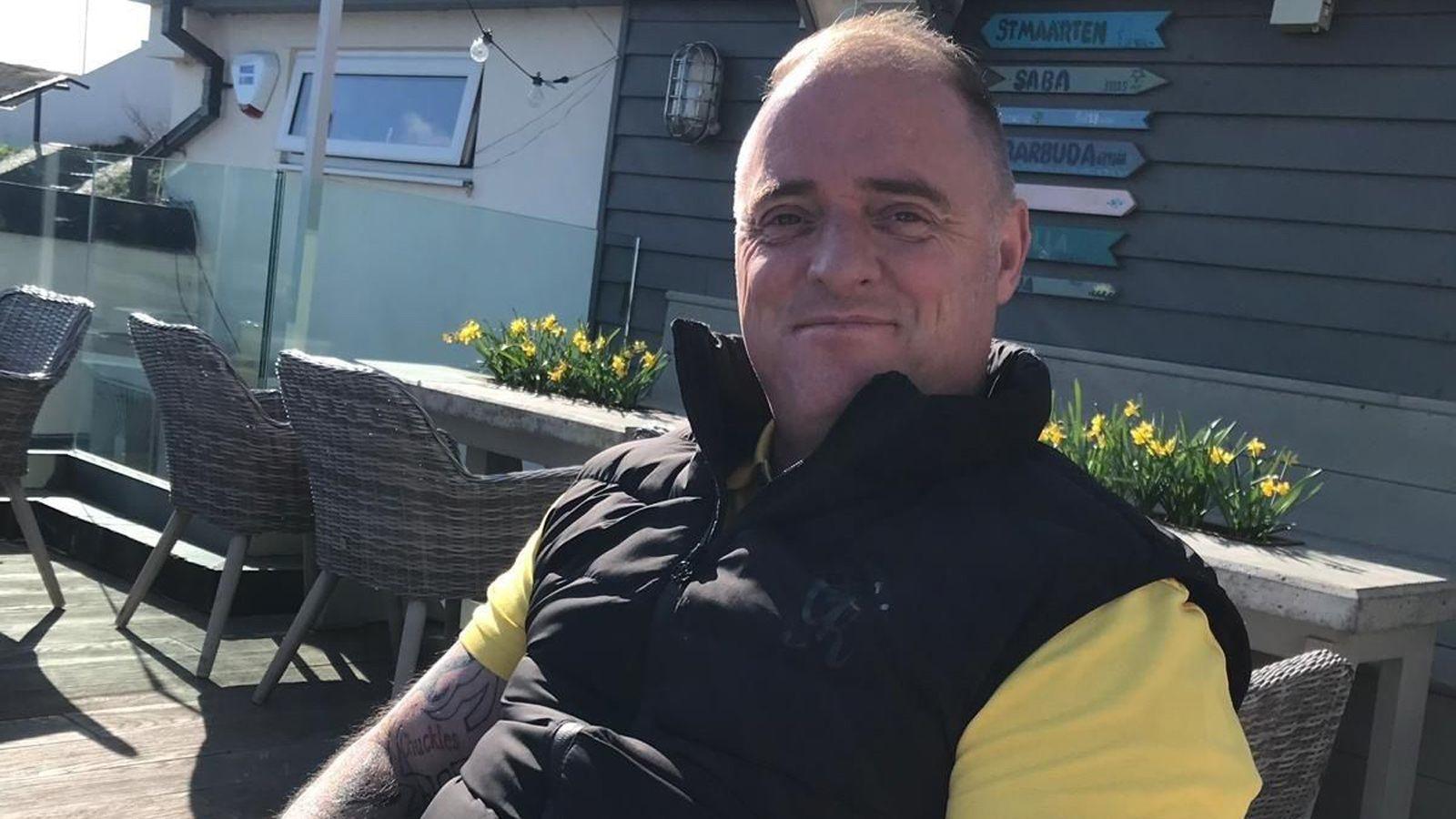 A smiling man with receding hair sits in a wicker garden chair. He has a black gilet over a yellow t-shirt. Behind him are rectangular, pointed wooden signs with the names of foreign places. They are attached to the dark wood cladding of a single-storey building.