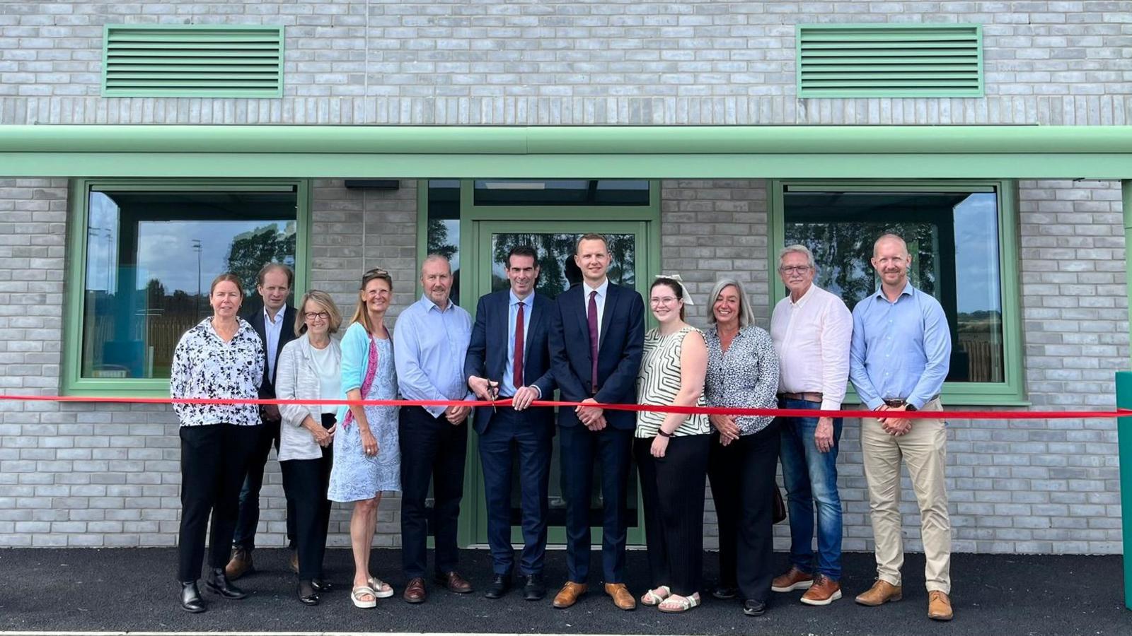 Men and women standing in front of a brick building with one of the men about to cut a red ribbon that is stretched out in front of them.