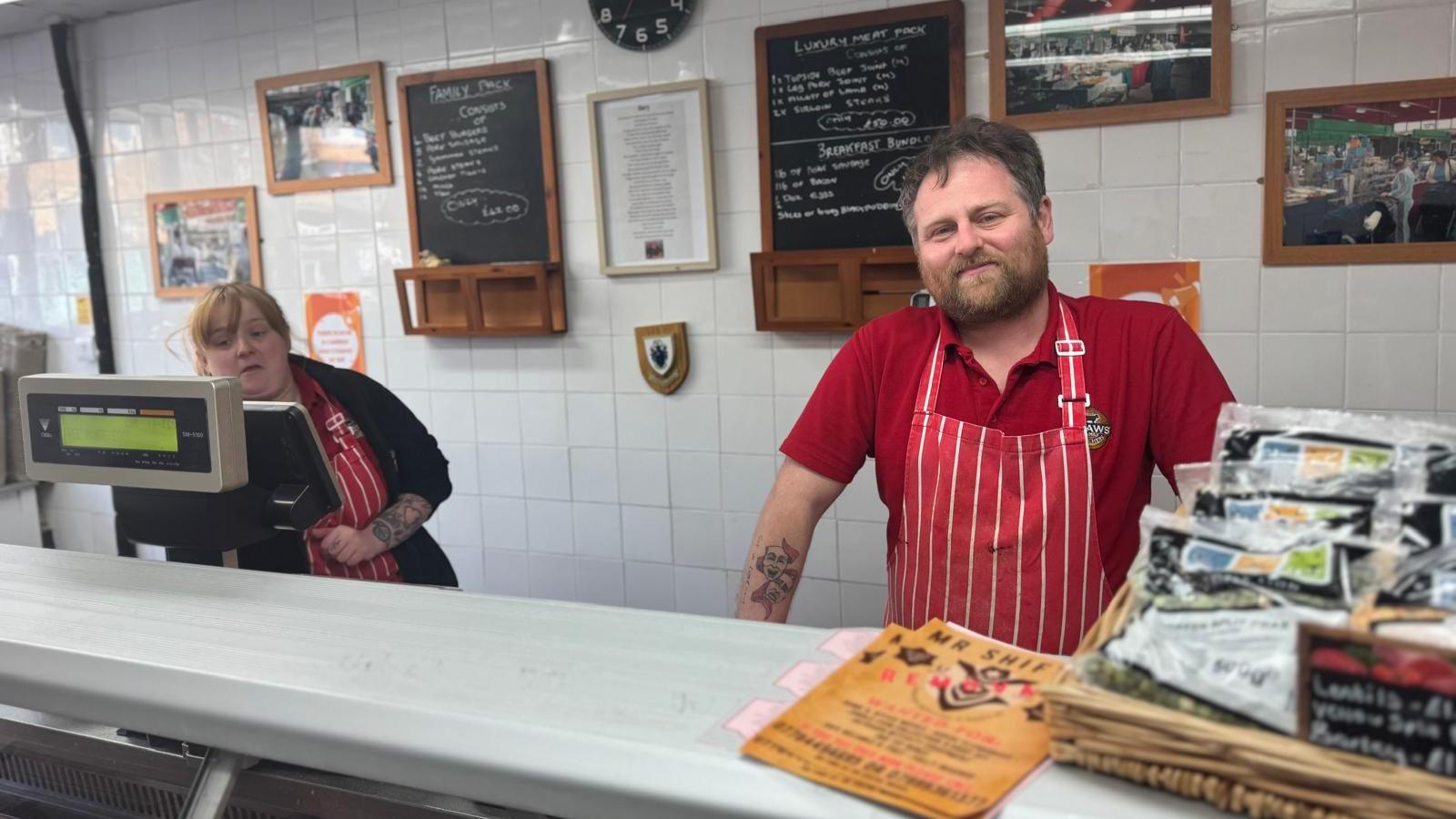 Danny Shaw, in a red polo-neck t-shirt with a red and white butcher's apron, stands behind the counter of his butcher's shop. Another member of staff, a woman in a black cardigan and also wearing an apron, is to the left