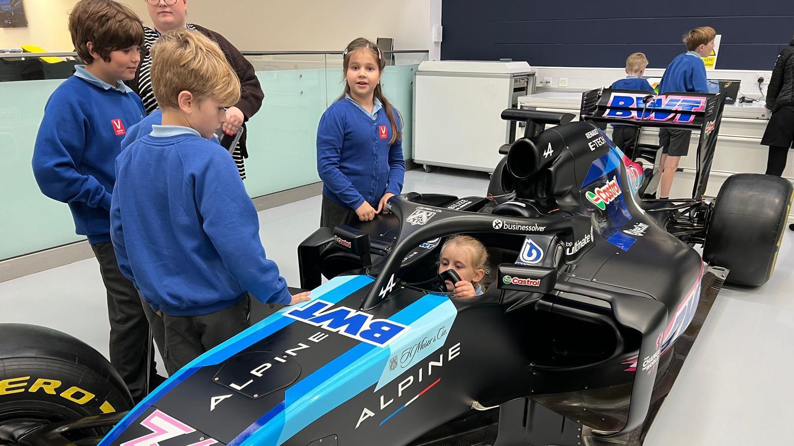 A young girl sits in a black formula one car holding the wheel. Her classmates are crowding around her. They all have blue sweatshirts on.