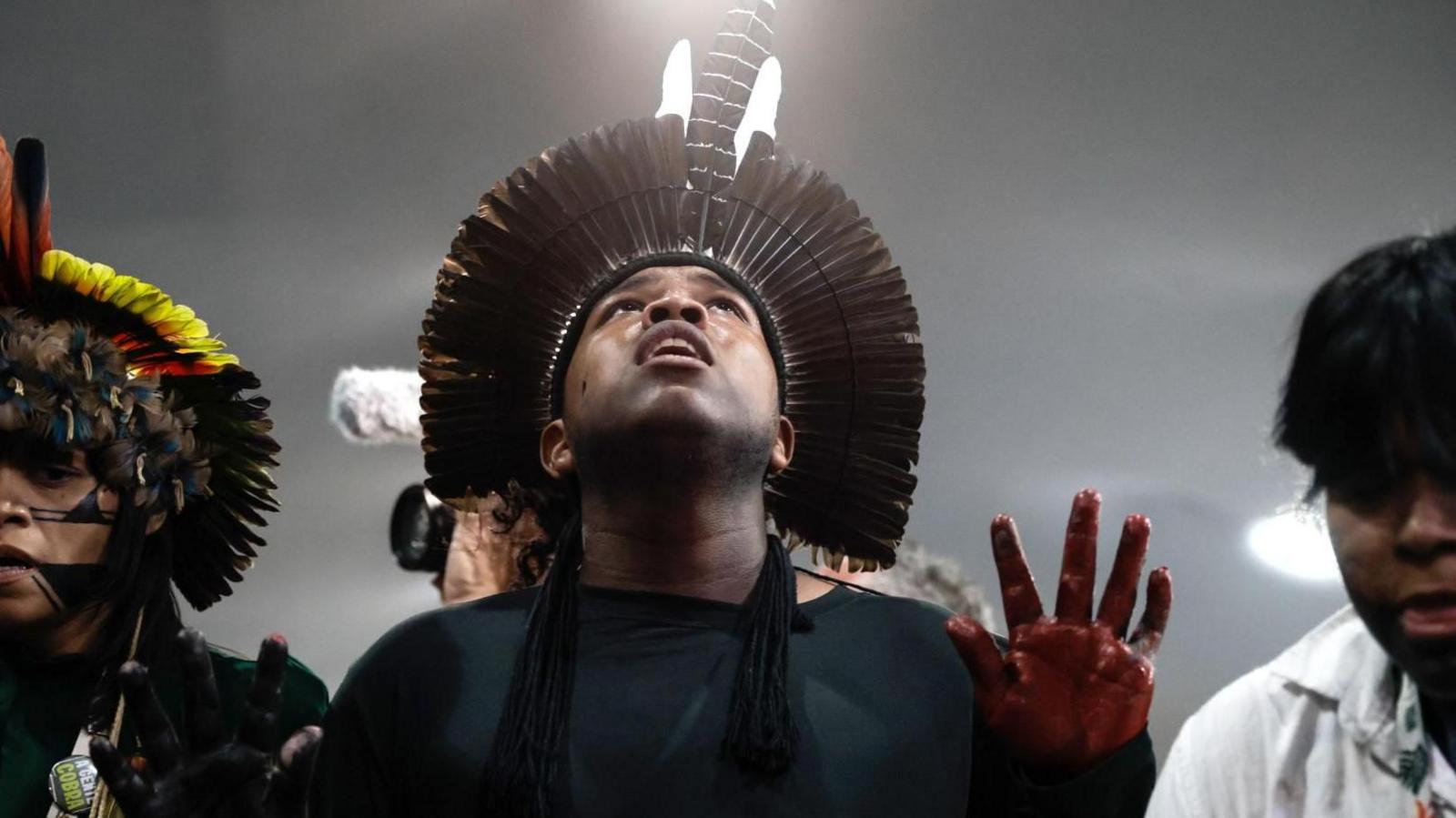 Indigenous people at a protest. Man in centre wears a headpiece made of features and red dye or ink on fingers