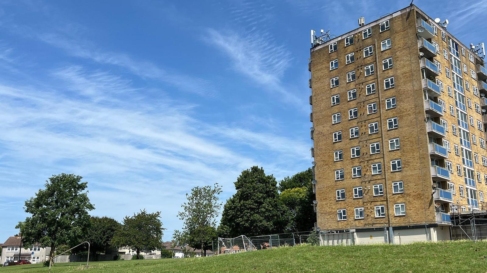 A red brick tower block with grassy park in front. A blue sky above. Some trees around too and a residential area in the distance.