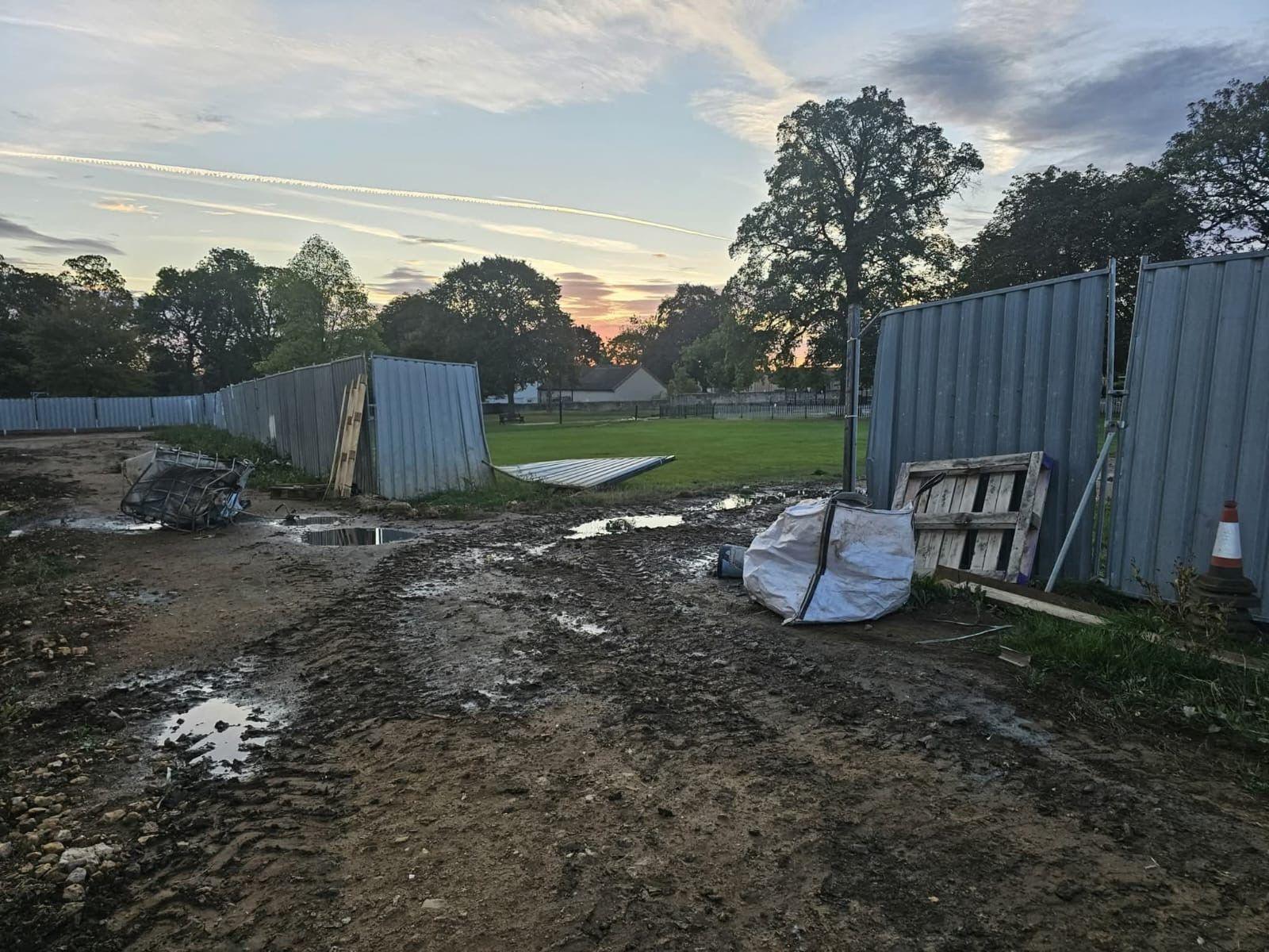 There is a building site with a large fence smashed down and a grass field churned up with mud