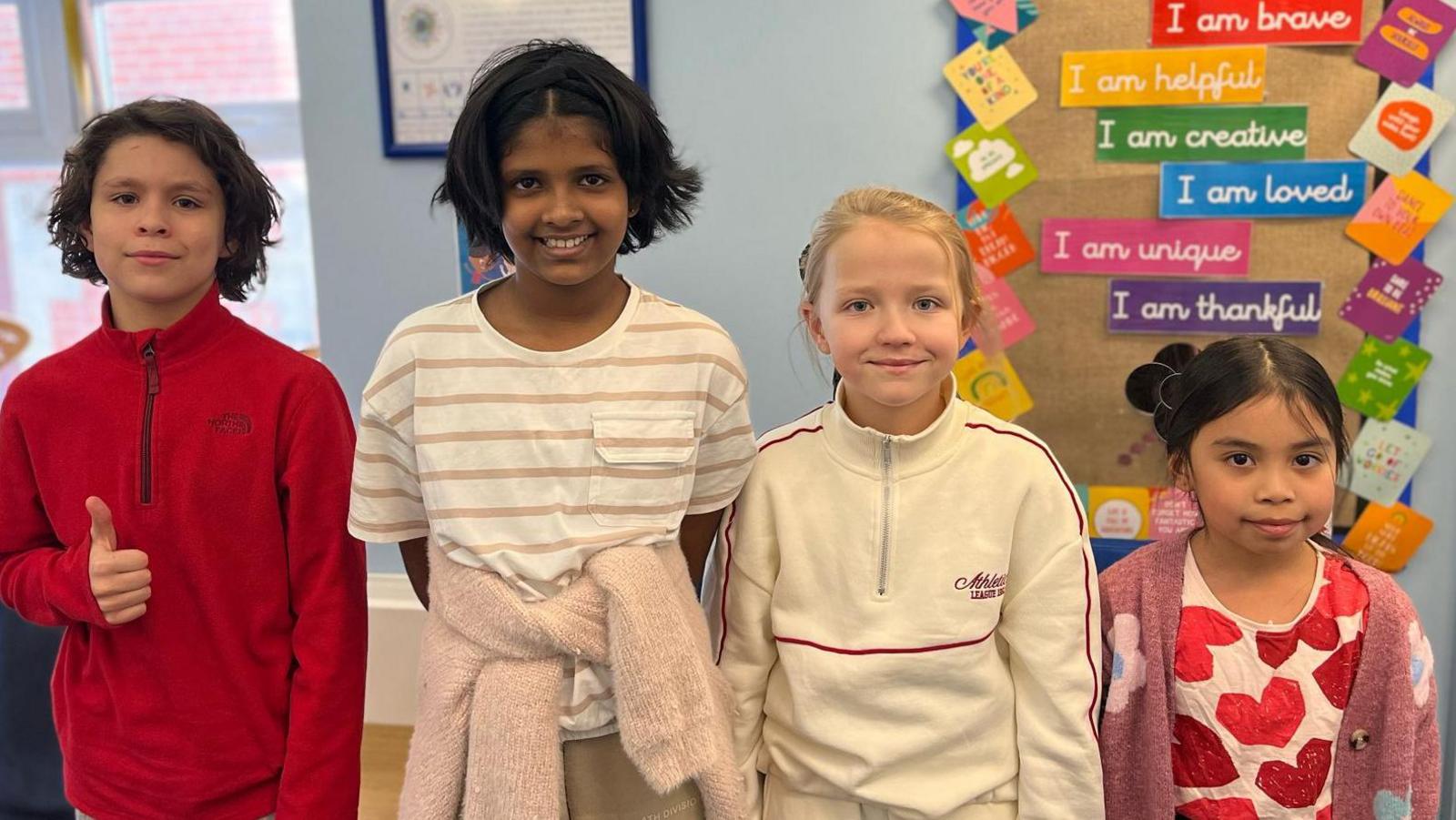 Four schoolchildren smile at the camera in a room at their school, with one giving the thumbs up.