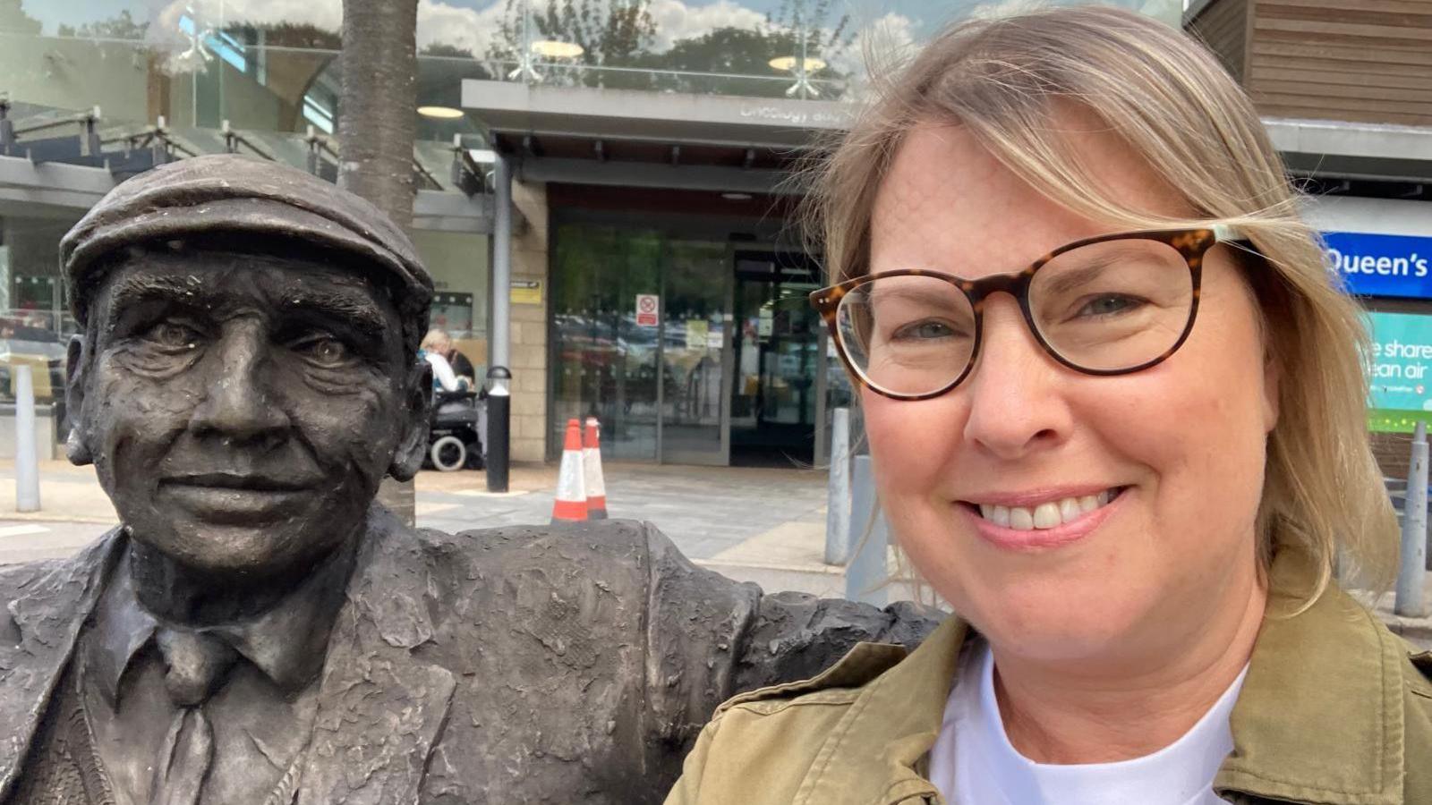 Woman taking a selfie beside a seated bronze statue outside a modern building with glass walls.