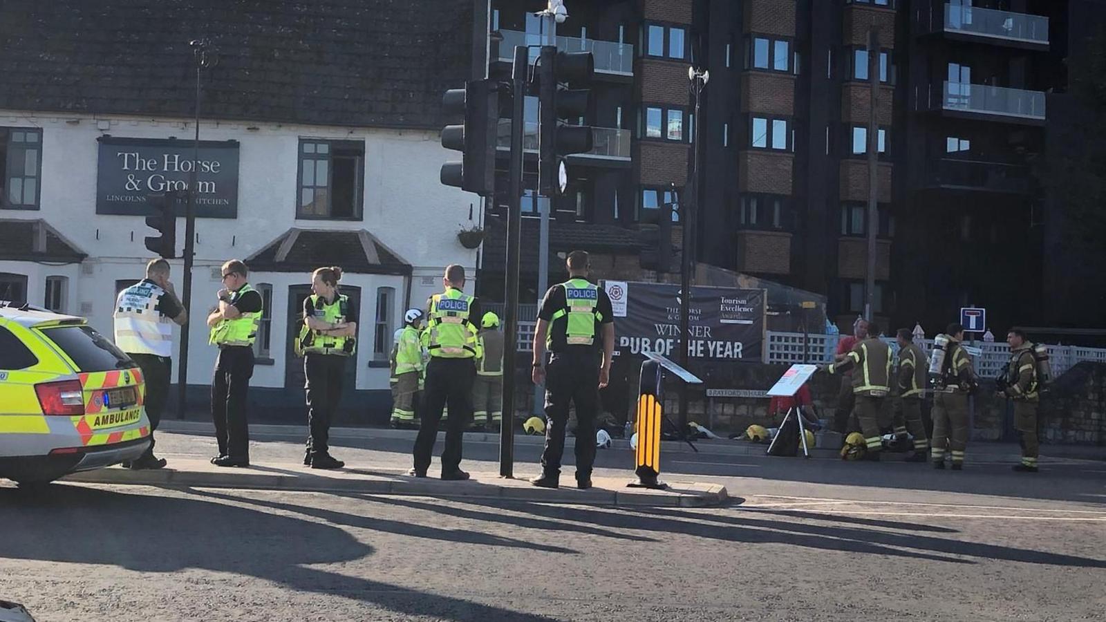 A street scene with a group of police officers and firefighters gathered near a building. An ambulance is parked on the left. In the background, a sign for "The Horse & Groom" is visible on a white building.