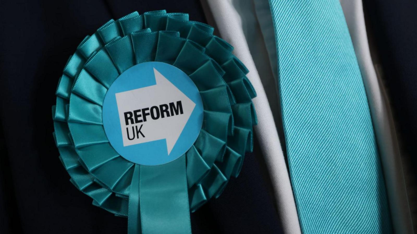 A close up of a turquoise coloured rosette with the words reform UK written in the centre. The rosette is pinned to the lapel of a black suit jacket being worn with a white shirt and turquoise tie.