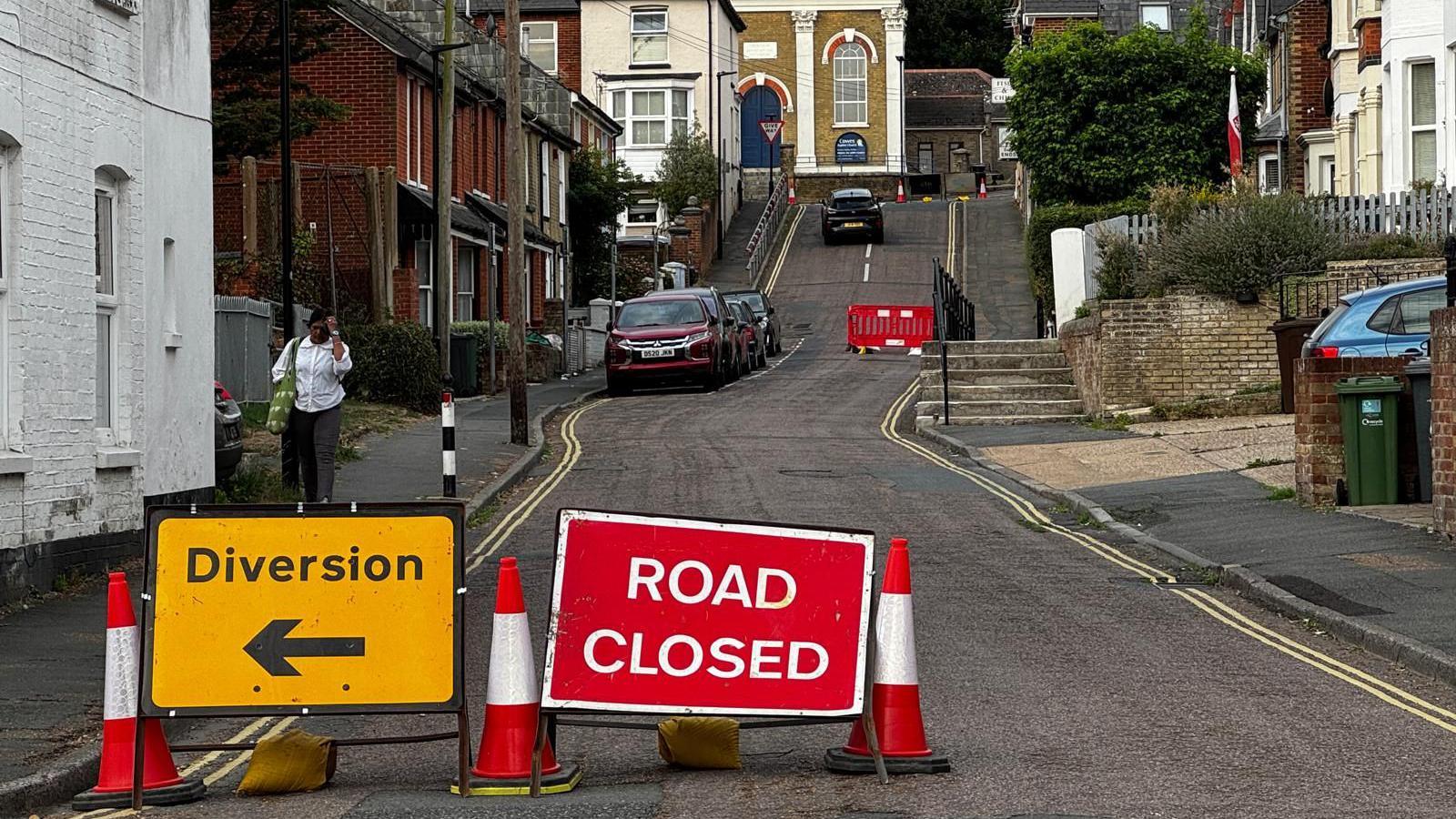 A residential street with the area cordoned off with a handful of cars parked on the left side of the road. A yellow diversion sign is pictured pointing left on the left hand side of the picture and a red ROAD CLOSED sign is in the centre of the picture.
