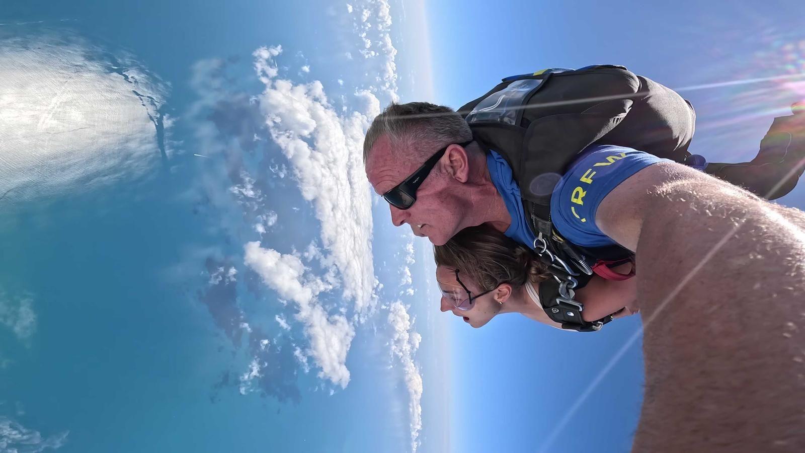 Lauren is sky diving with clear blue water under her. Her sky diving instructor is on her back and he is holding the camera as the wind pushes his cheeks back. They are harnessed together. 