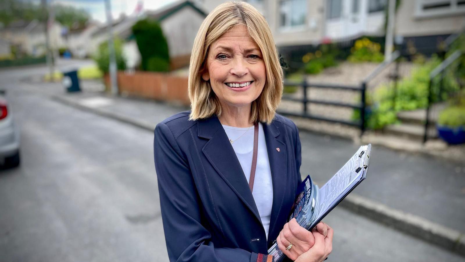 Diana Armstrong holds a clip board, she has light brown hair and wears a white t-shirt under a navy blazer