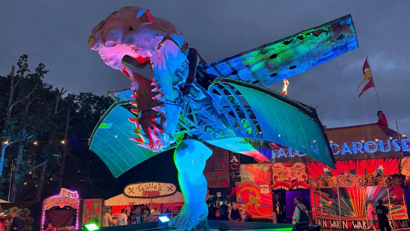 A monster's head that has a lower jaw similar to that of a T. rex with wings and a human leg. Lighted up at night in blue, green and pink colours.