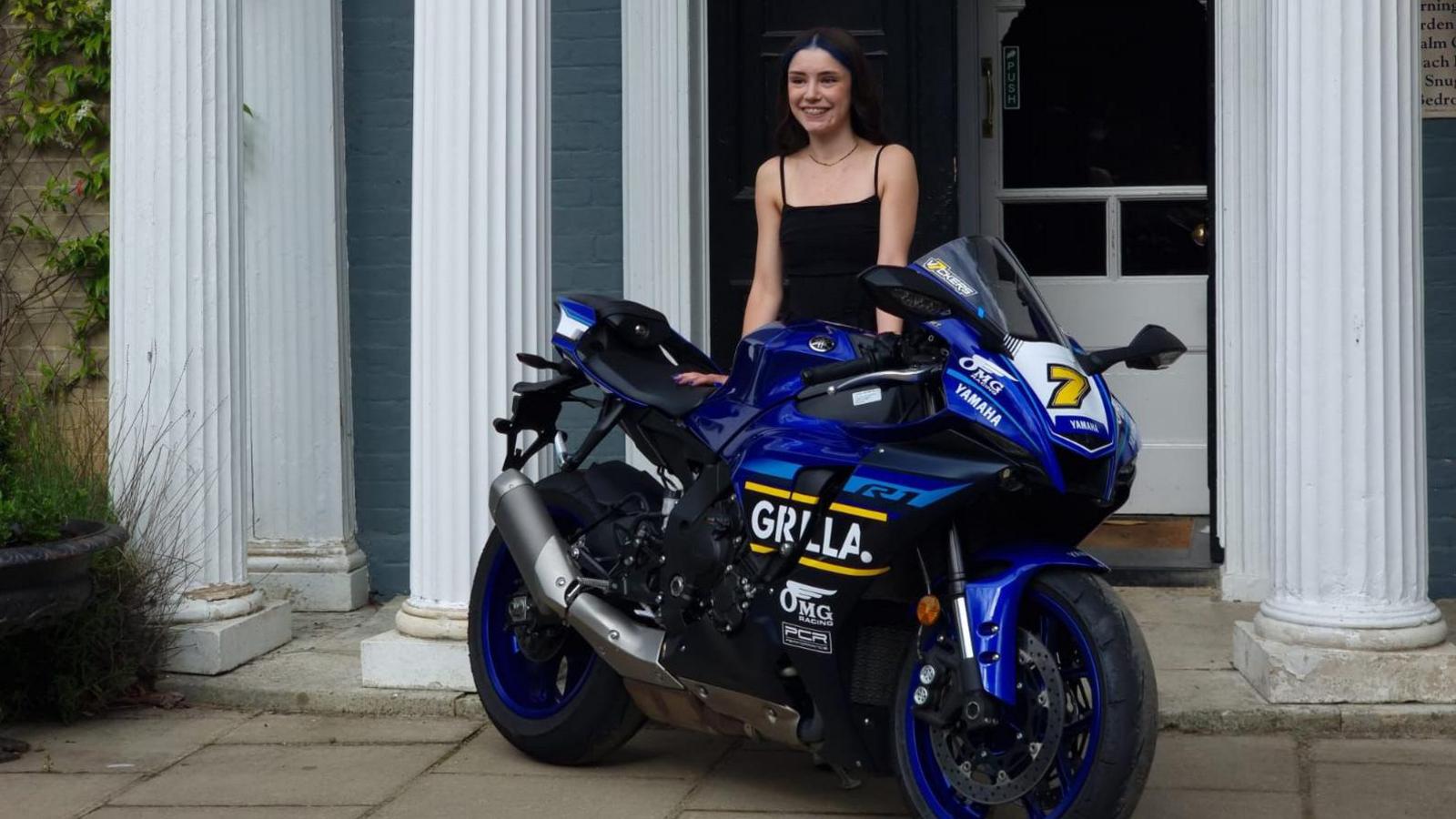 Scarlett Alexander in a black dress standing in front of Caistor Hall Hotel with a blue OMG replica superbike