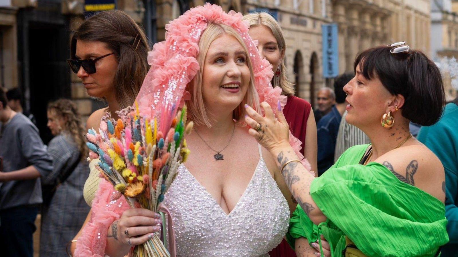 A smiling woman in a bridal dress holds a bunch of colourful flowers as a friend in a green top helps adjust a pink head decoration. In the background other well wishers can be seen