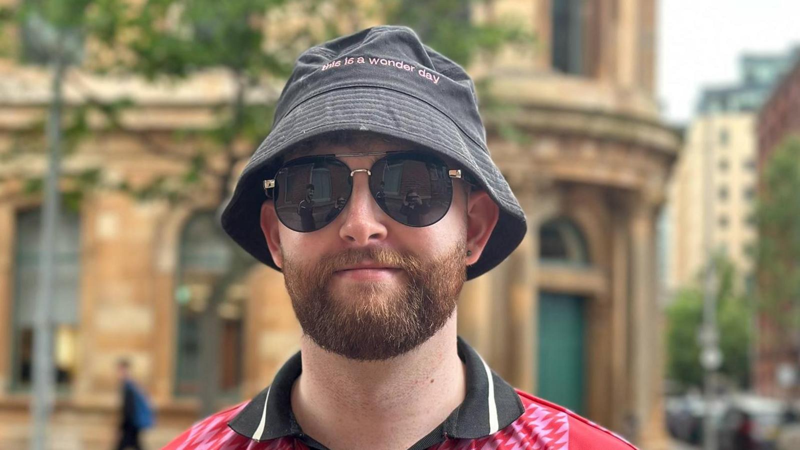 A young man with a ginger beard wearing a black bucket hat, sunglasses and a red and black collared top.