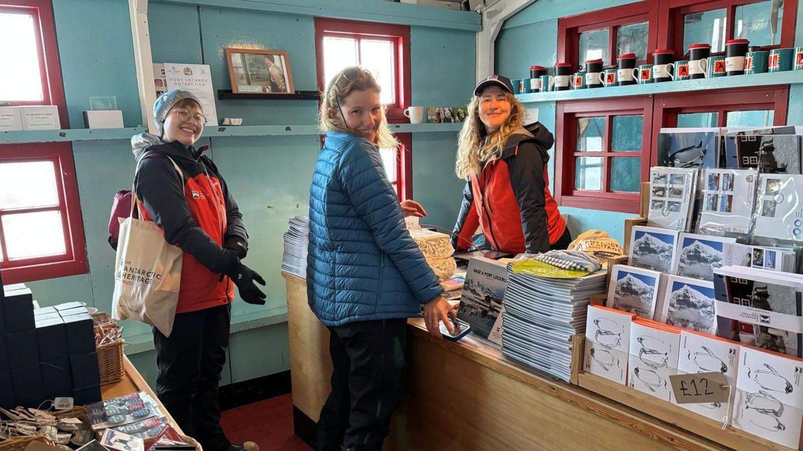 A picture showing Dale Ellis with two female team mates. Dale is stood behind a desk in the shop with her team mates in front. There are postcards and cups on display. 