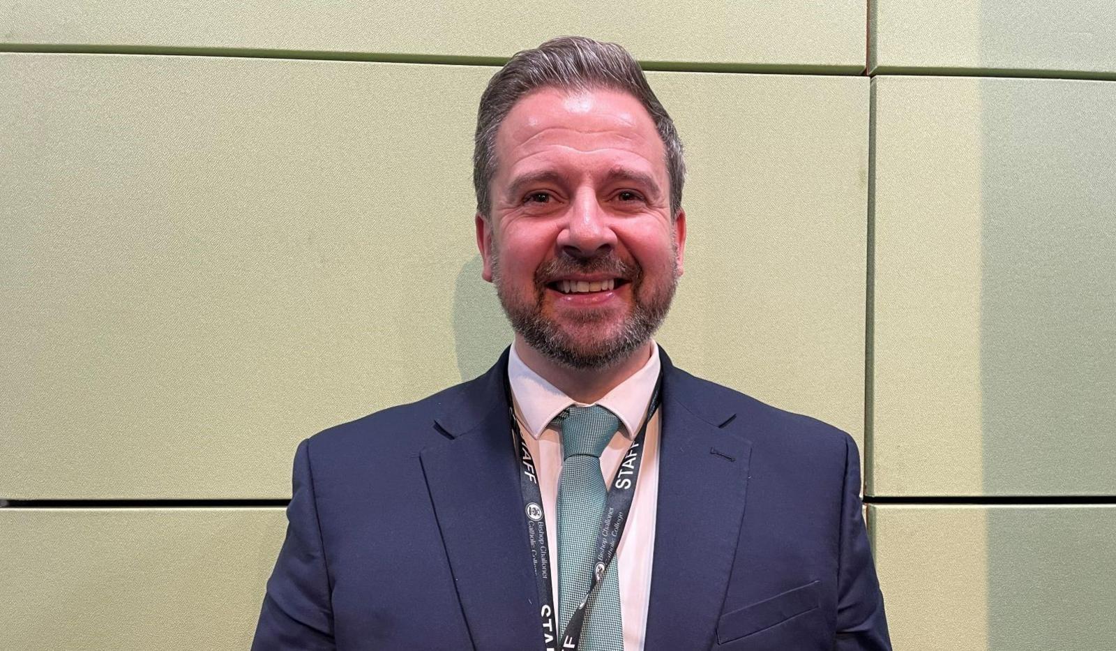 A man stands against a cream-coloured wall. He has grey hair and a grey beard. He is wearing a dark blue suit jacket, a pale pink shirt, and a pale green tie.