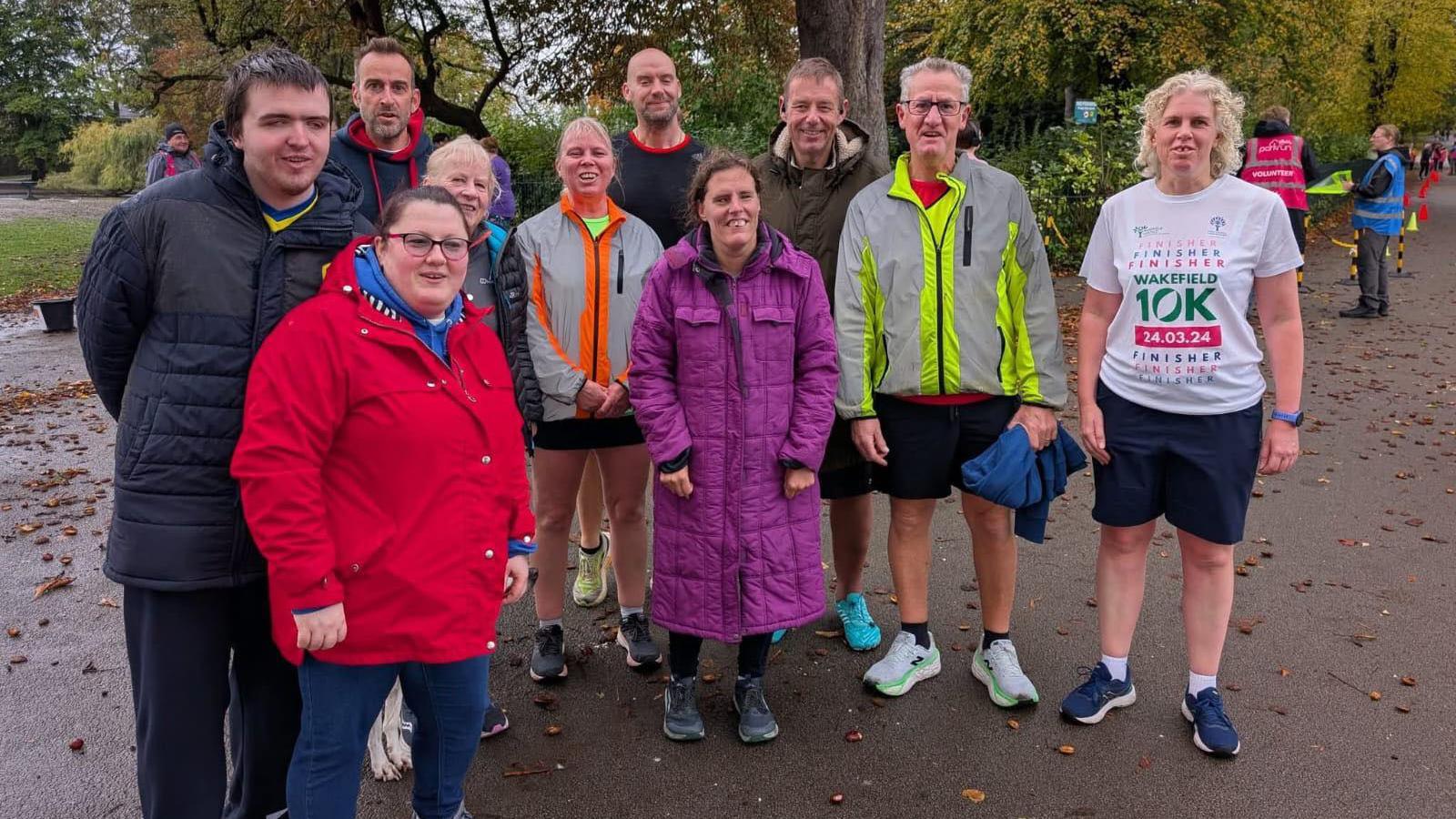A large group of people huddle together smiling for the camera. Some are dressed in winter coats, others wear running clothes. Behind them are tall trees and leaves are scattered along the path they stand