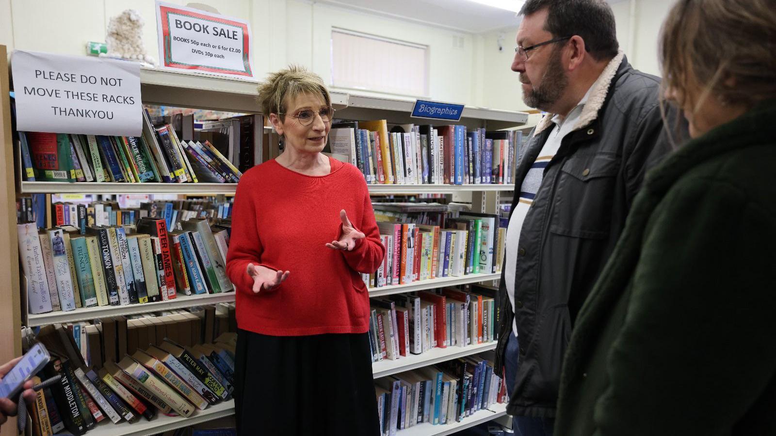 Jennie wearing a red jumper and black skirt speaking to Harper- wearing a black jacket and a white T-shirt- standing in front of book racks 