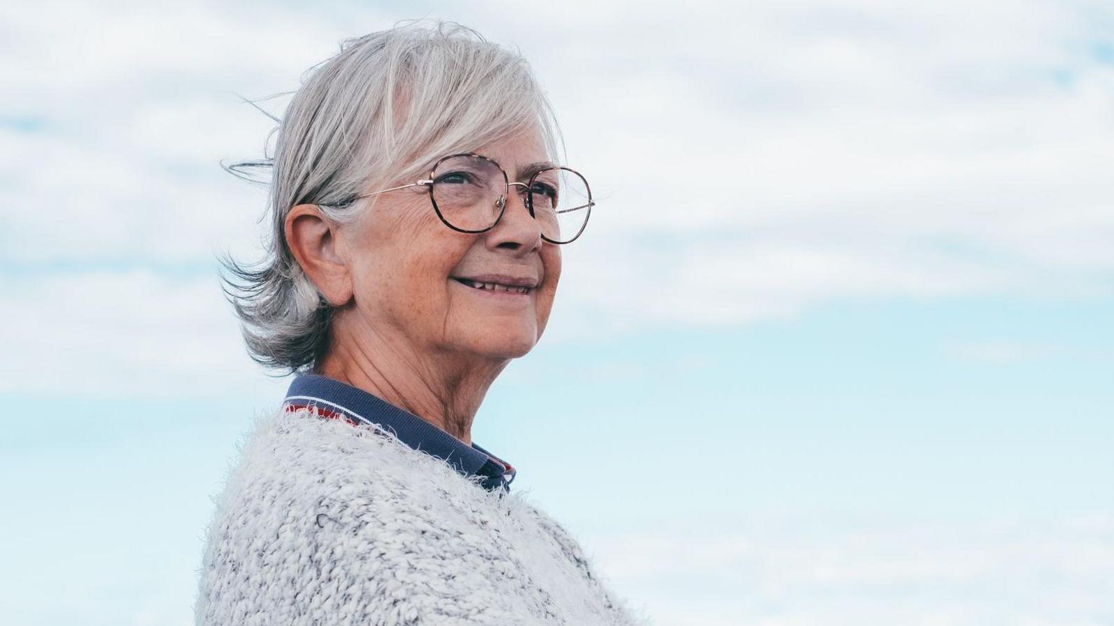 Smiling elderly white-haired lady stands by the sea. She wears glasses and a fluffy grey cardigan and stands with her arms crossed, looking off camera.