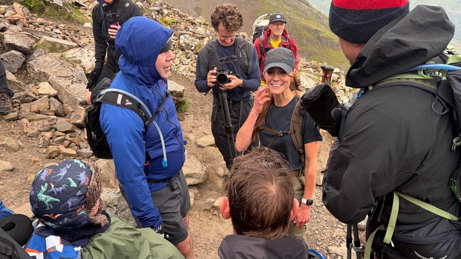 Catherine, Princess of Wales, speaks to a boy in a wheelchair and a group of men while on a mountain. She wears a grey cap and black t-shirt.