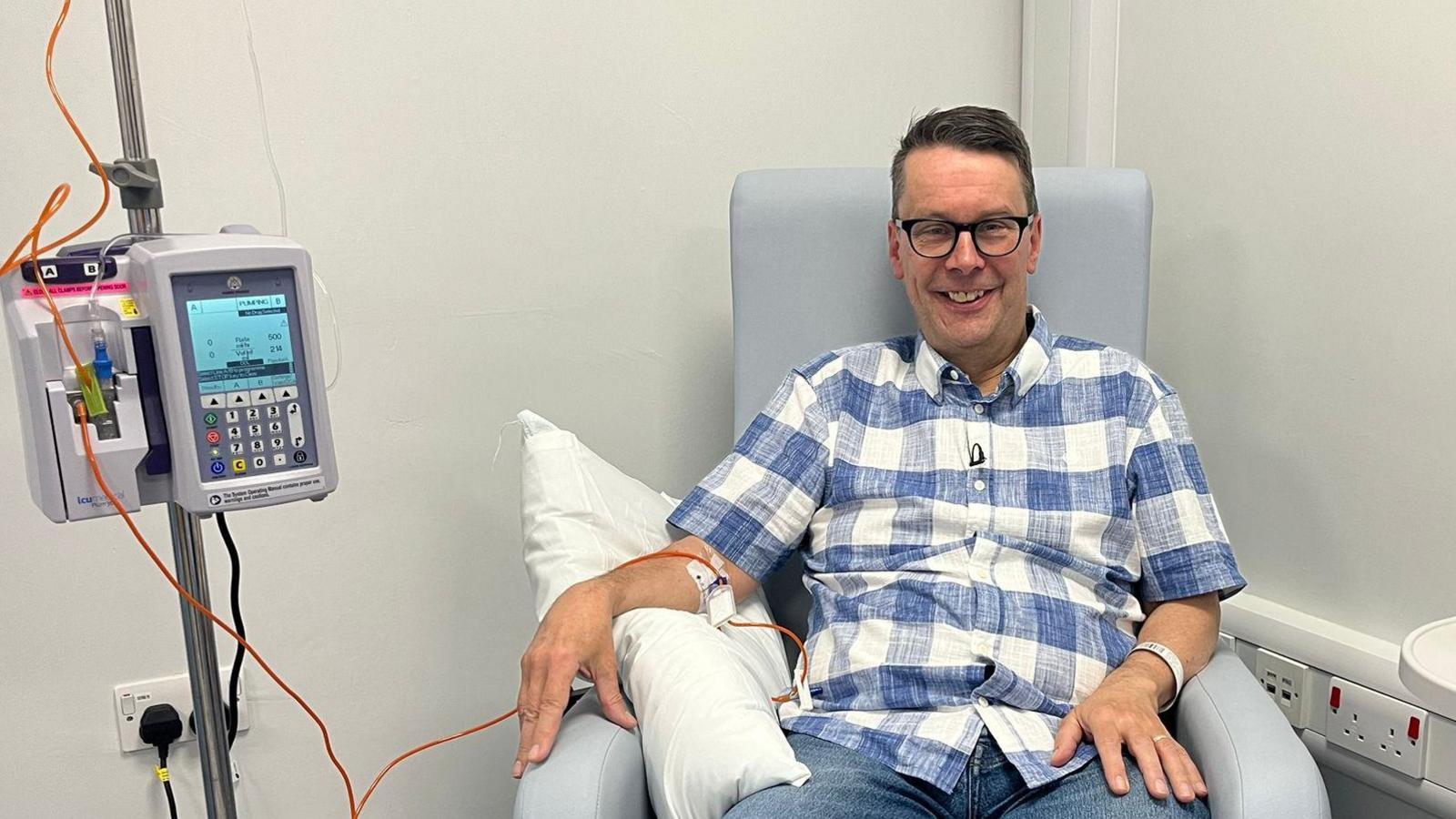 Paul wearing a blue and white checked shirt is sat in a chair, bare arm is resting on a cushion while an orange tube connects a cannula to a pump.