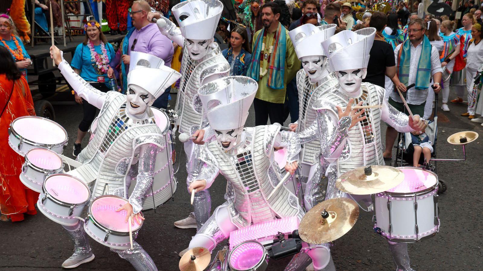 four people dressed in mirror clothing. Their faces are painted white with black eyemakeup. Thy are holding drum sets around their waist. 