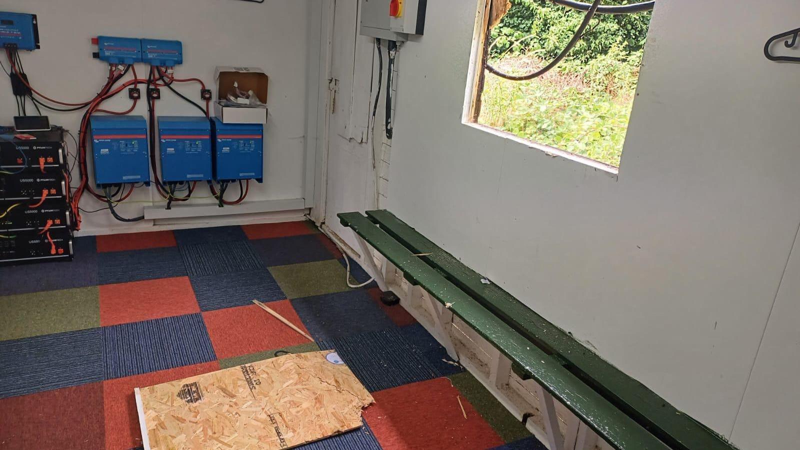 A room with a broken window and a wooden board, which has fallen on a checked carpet.