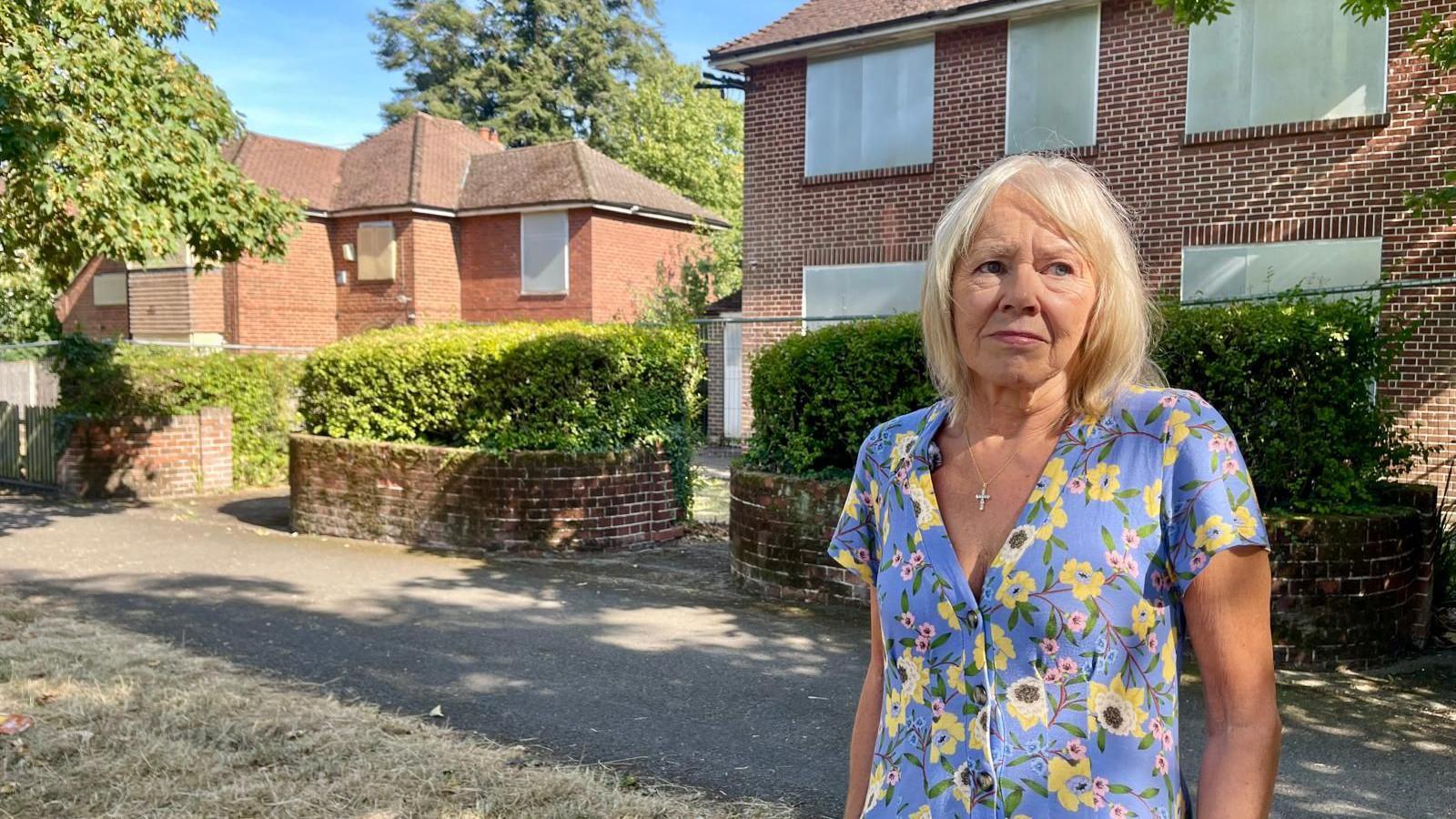 A woman with blonde hair and a flowery blue dress stands outside two large houses with hedges and a driveway. The windows of both houses are bordered up and the gardens surrounded by metal fencing.