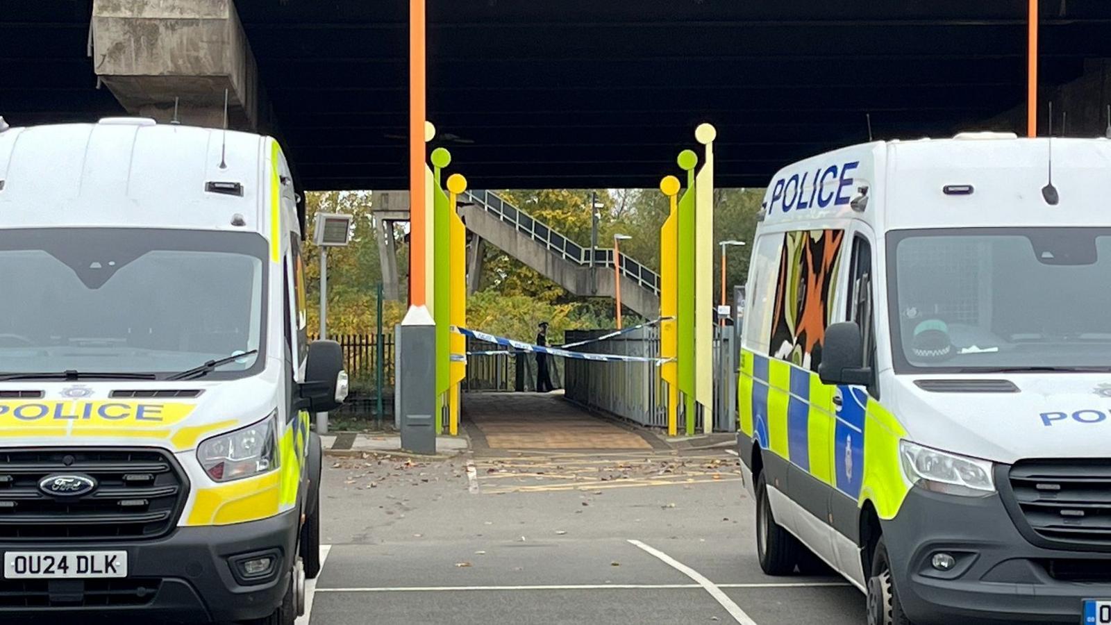 Two police vans parked in front of a path that is blocked off with two strips of police tape. The police vans have blue and yellow squares on the side and the words "police" on them. Behind the tape, a paved path leads towards a concrete stairway going upwards.
