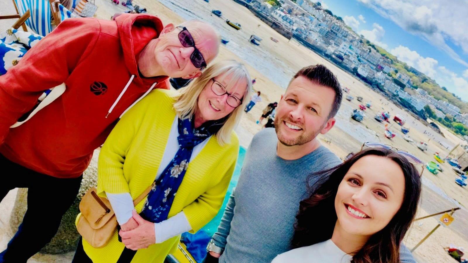 A group photo of four people on a beach in St Ives. From left to right, there is a man in a red hoodie with black sunglasses, a blonde woman with glasses wearing a yellow cardigan and a blue scarf, a man wearing a grey sweater, and dark haired woman with sunglasses propped on top of her head. 