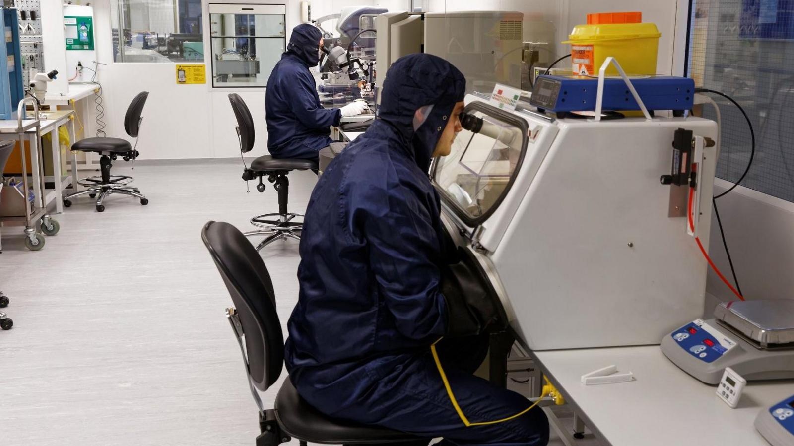 Two scientists in a lab working on building the Leonardo sensors in the Space Lab at Leonardo in Southampton. They are wearing blue fitted costumers. They are looking into microscopes.
