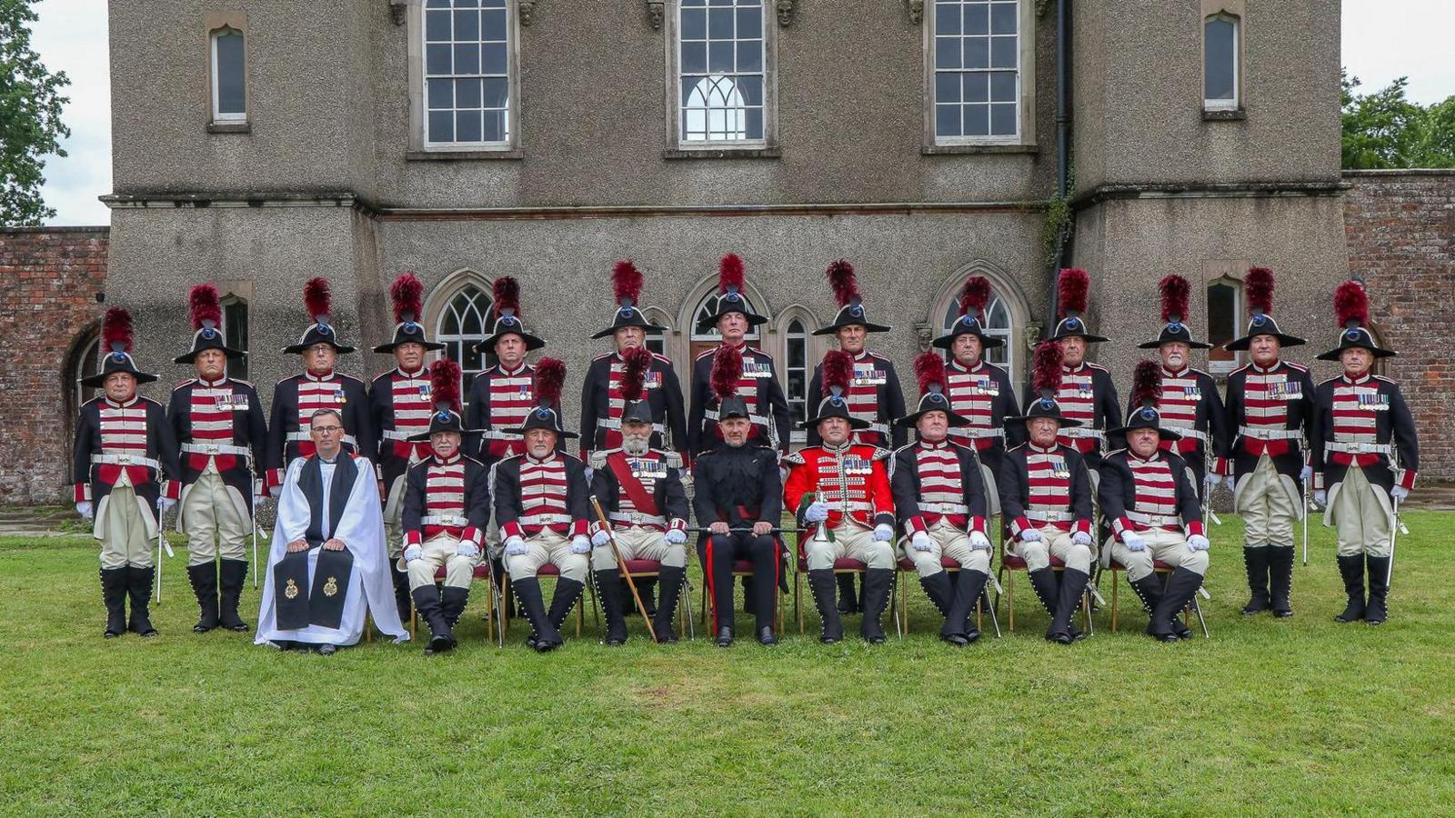 20 men in full ceremonial dress, including military style jackets and hats with feather plumes. The uniforms are navy, with white and red stripes across the front. They are wearing black boots and have swords on their sides.