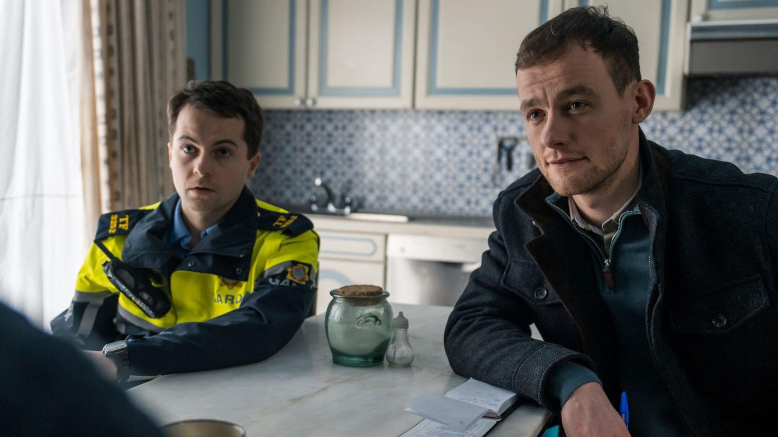 A still of Alex Murphy on the left wearing a police uniform. Sitting beside him is Alan Mahon wearing a dark coat and a green half-zip jumper. The two men are sitting at a kitchen table, talking to someone who is off-screen. The cabinets behind them are white, with a patterned blue and white tile.