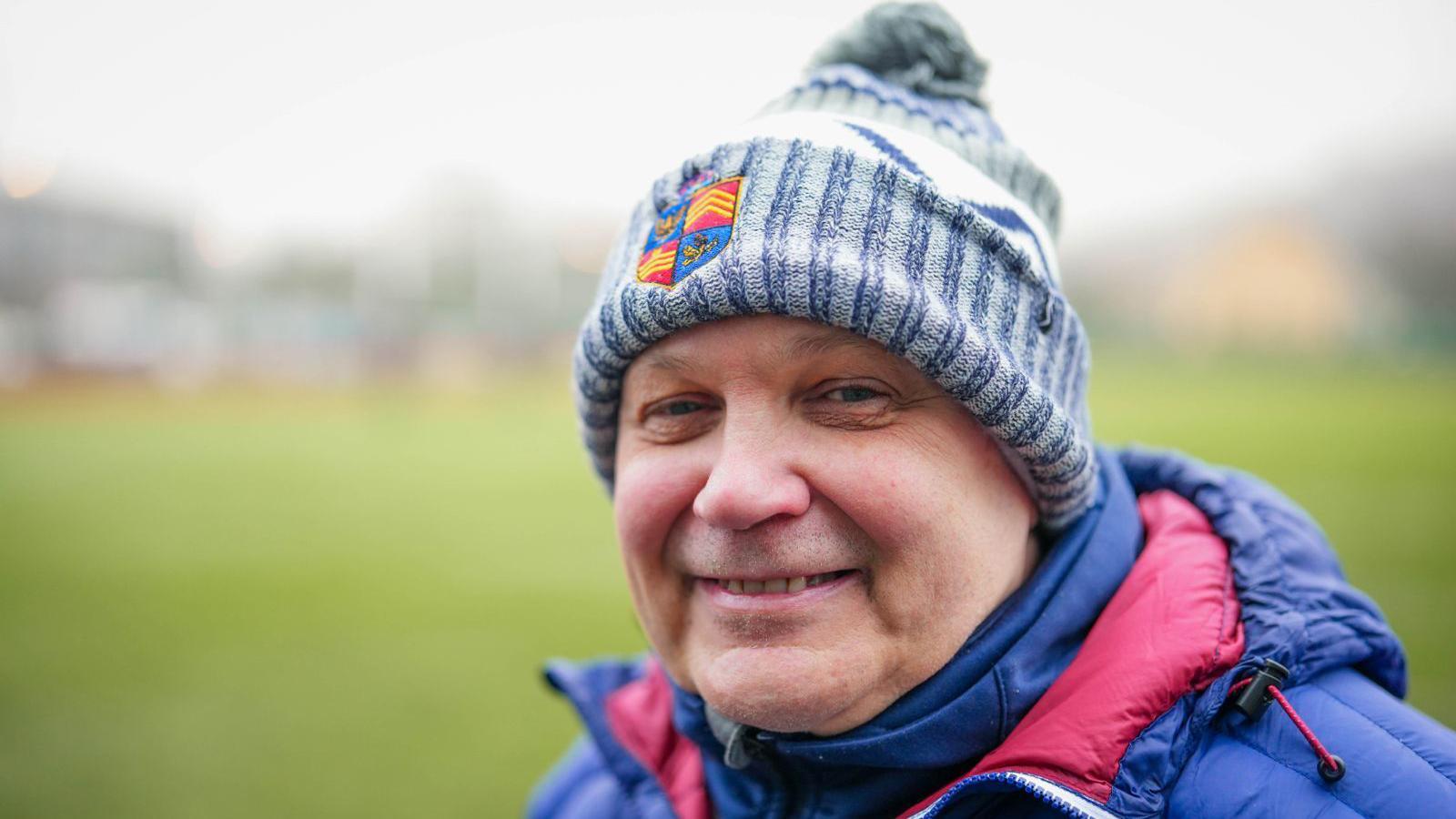 Darrell Williams smiling at the camera with the blurred rugby pitch in the background. He is wearing a blue and white bobble hat and a navy and red coat. 