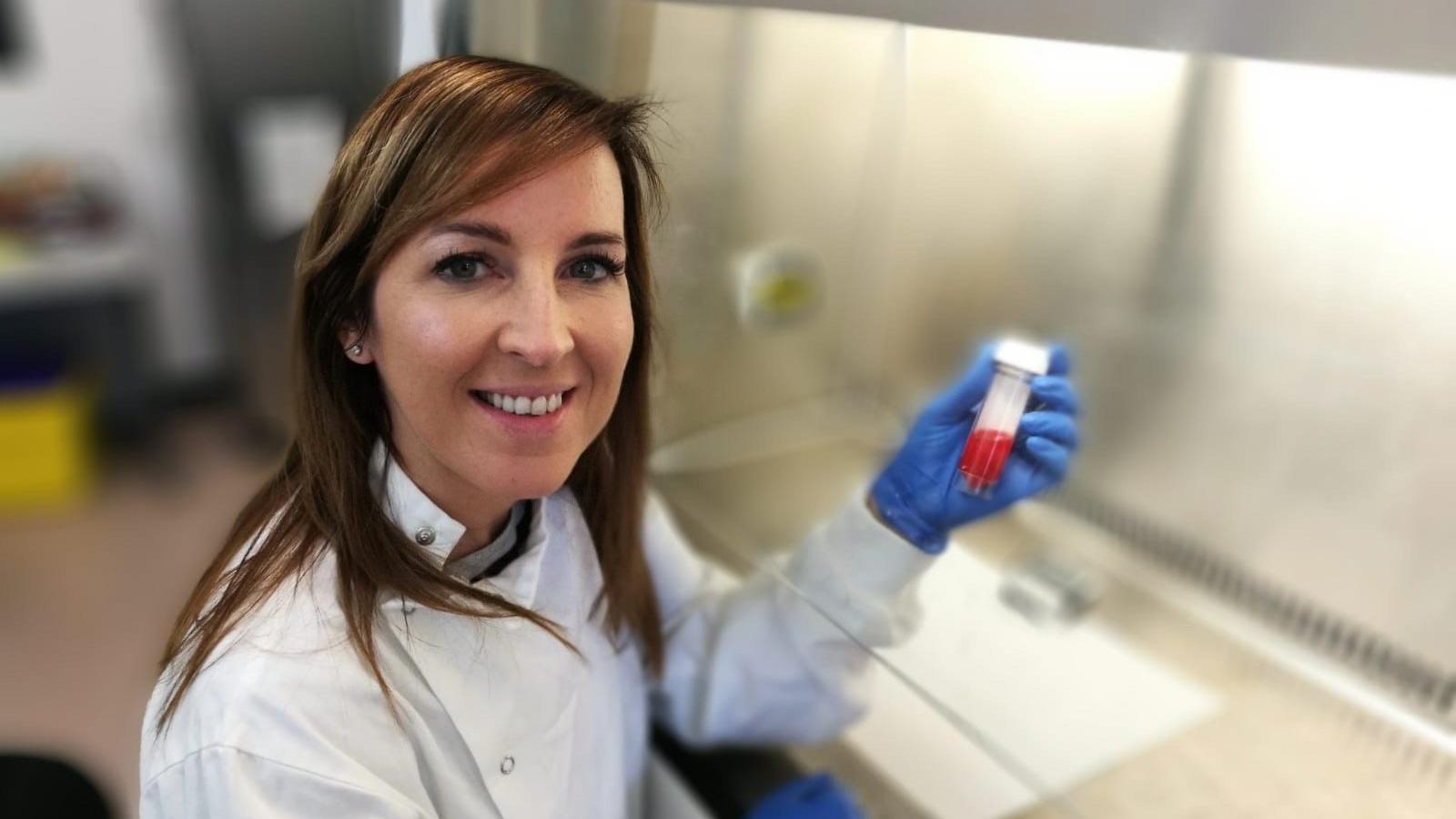 Trisha sitting smiling at the camera. She is wearing a white lab coat and blue gloves and is holding a vial with a red liquid in it.