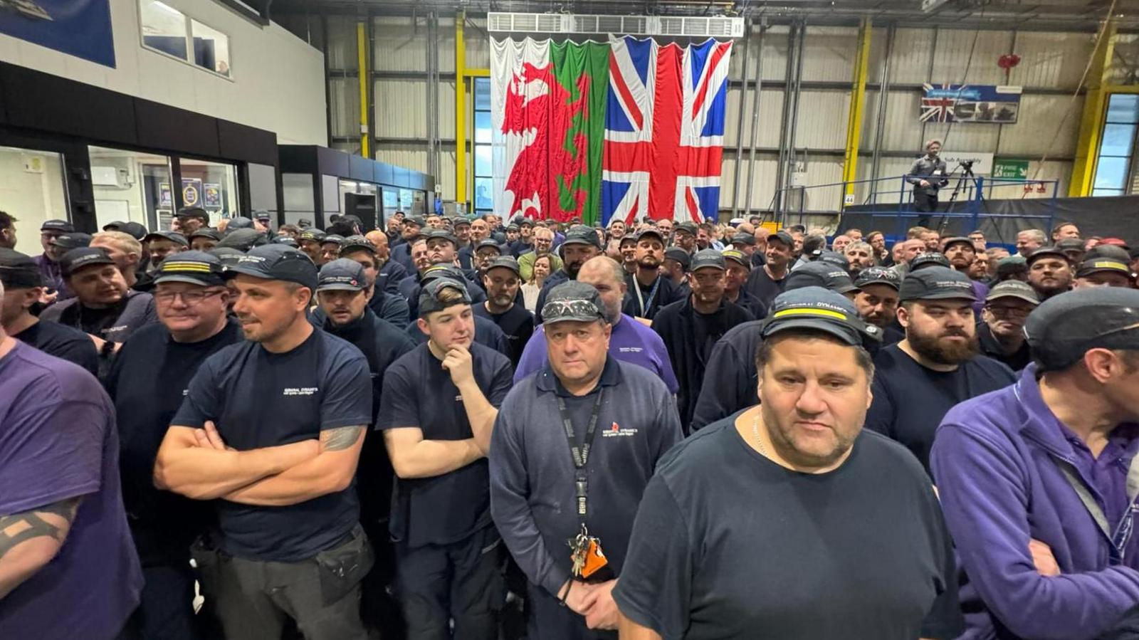 A large group of workers standing inside the Merthyr factory, with a large Welsh flag and Union flag on the wall at the back of the plant