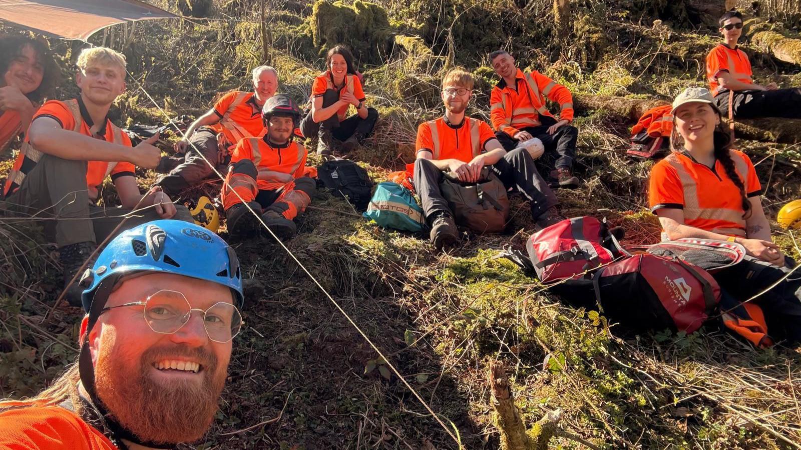 Workers in orange high vis strewn sat on a steep bank smiling for a group photo