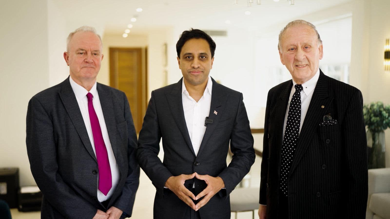 Three men in suits pose for a photo. Martin Griffiths is on the left, wearing a dark pink tie and charcoal suit. Ken Harrington is on the right with a black and white patterned tie and pocket square and black suit. In the middle of the image, Zia Yusuf stands with the fingertips of both of his hands together. He is wearing a black suit and a white shirt