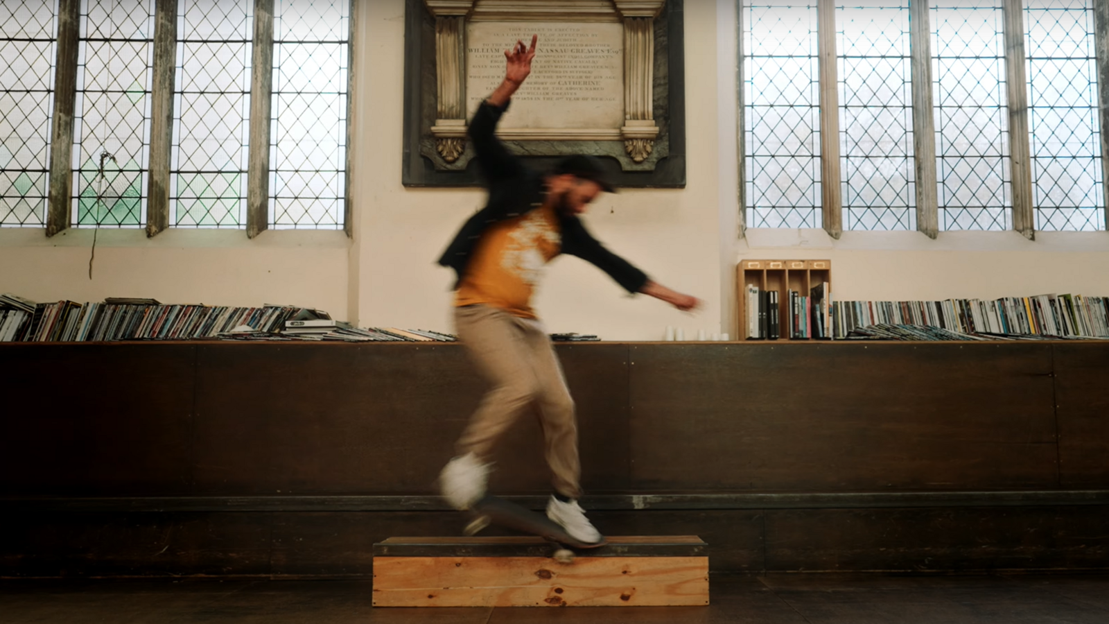 A man in motion, jumping on a skateboard. He is in an old church building, behind him there are books lining a shelf, large windows, and an old inscribed plaque.