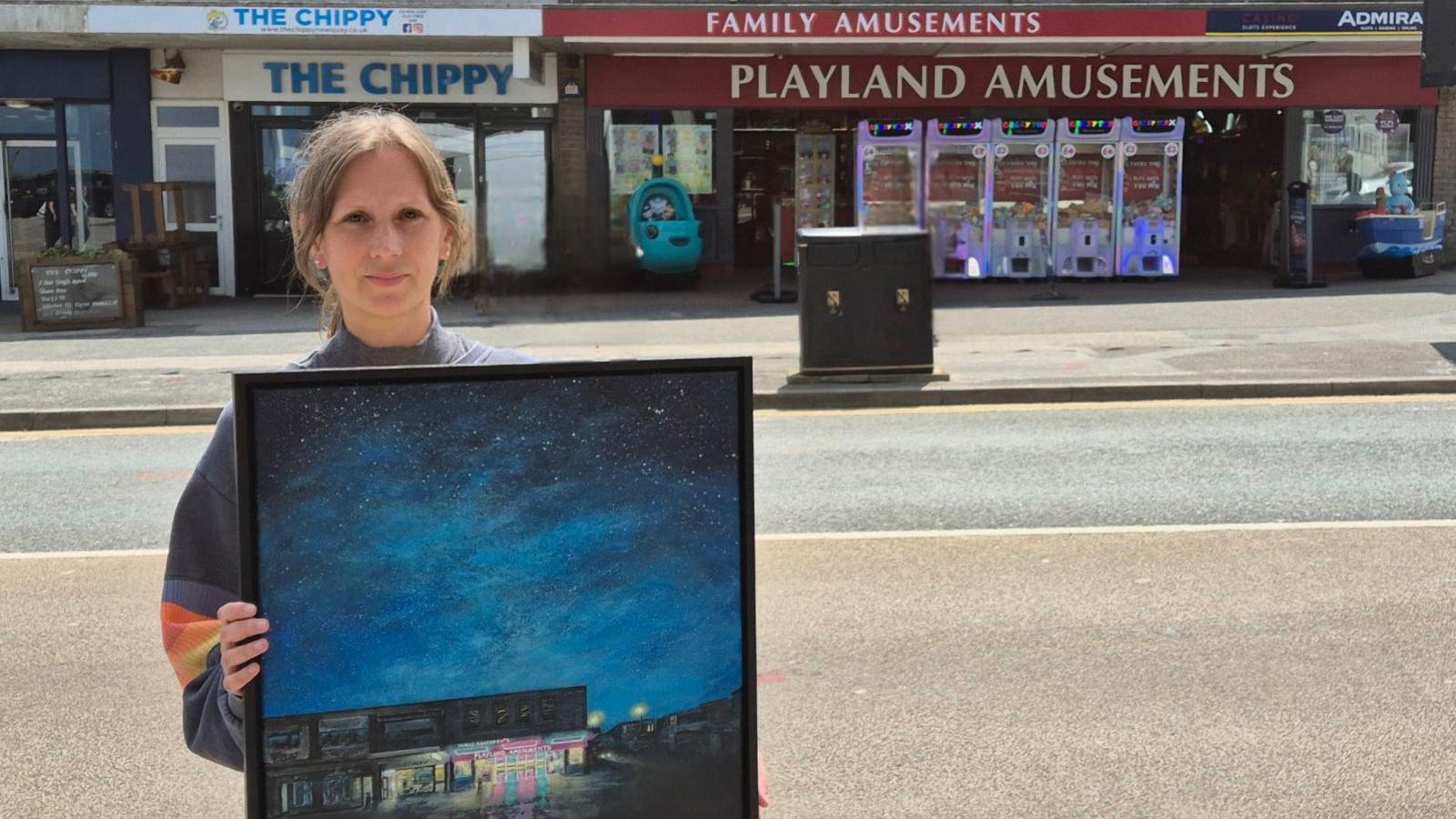 A picture of Dianne holding a picture of her blue painting outside of the arcade.