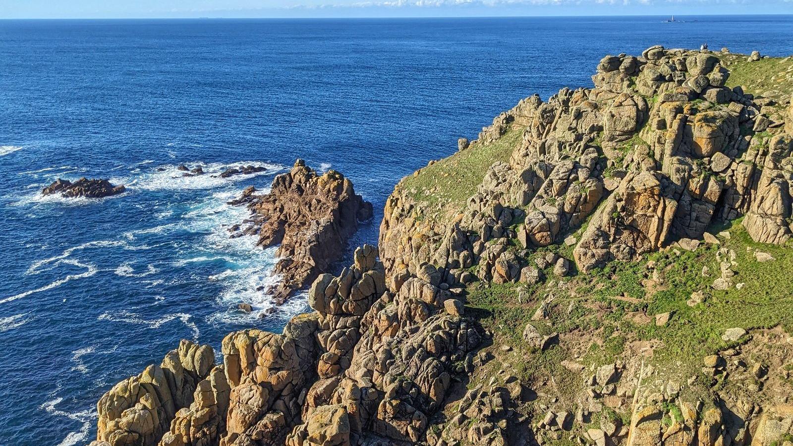 Jagged stones on a cliff face in Cornwall with the sea having a dark blue hue on a cloudy but sunny day.