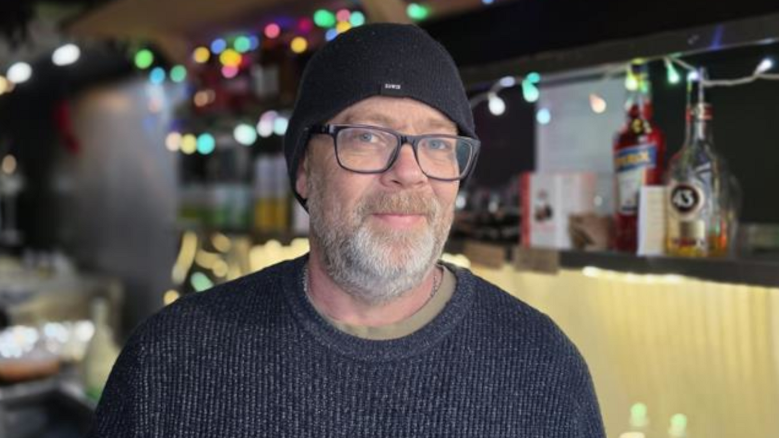 Hugo Malik, is standing inside his market stall, Churros for the People. He is wearing a black jumper and black beanie hat. He is looking directly at the camera and smiling.