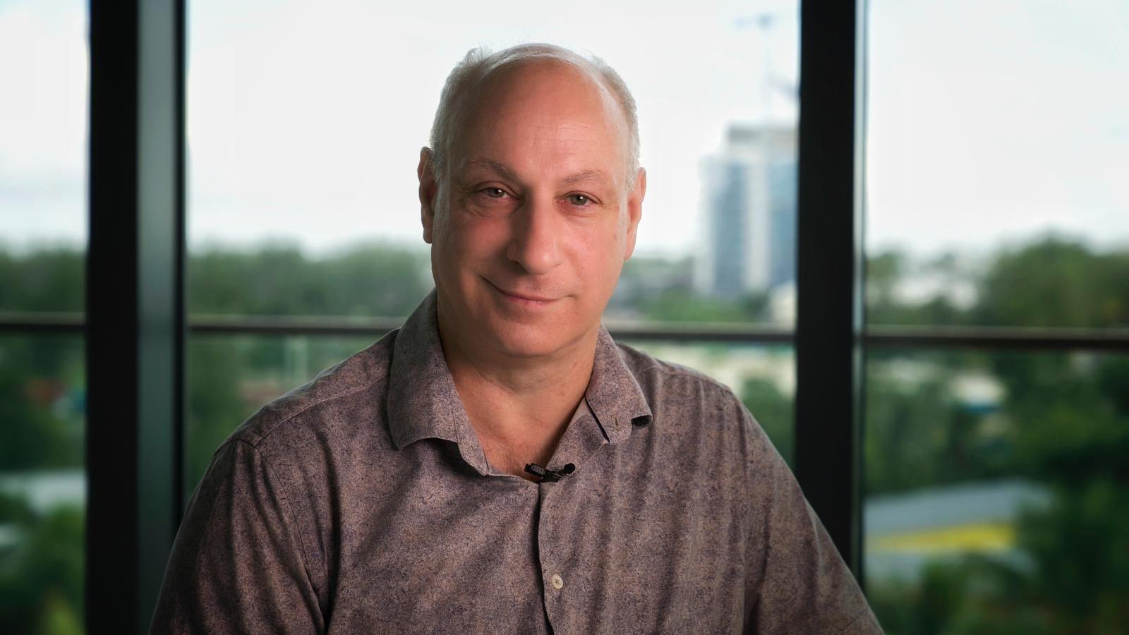 Adam Caller smiles at the camera from inside a building, sitting by a window
