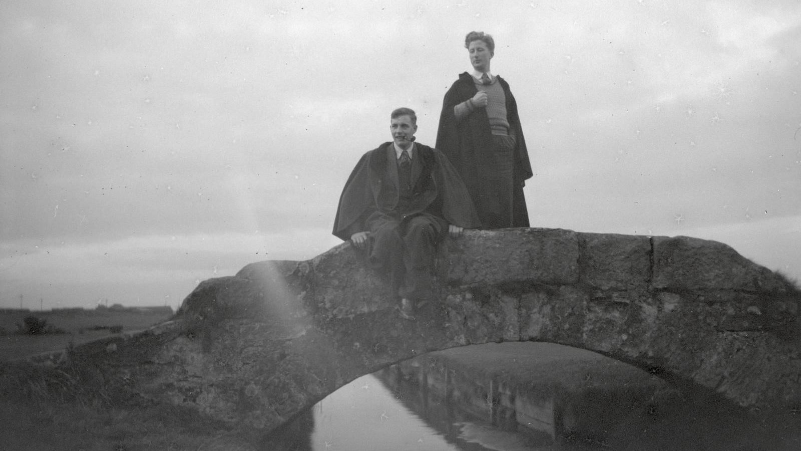 A black and white photograph of two young men on a small stone bridge over a river or canal, wearing ties with long back robes. One has a pipe in his mouth.