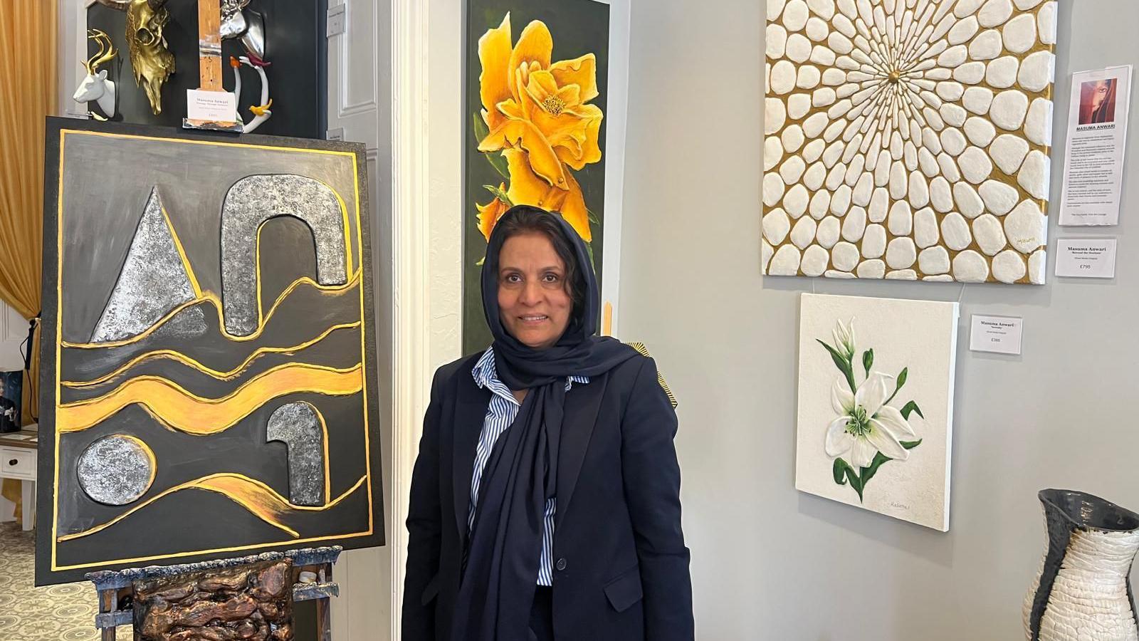 A woman in a blue blazer and headscarf with a striped blue shirt is standing in front six different pieces of artwork. They range from abstract shapes to flowers.